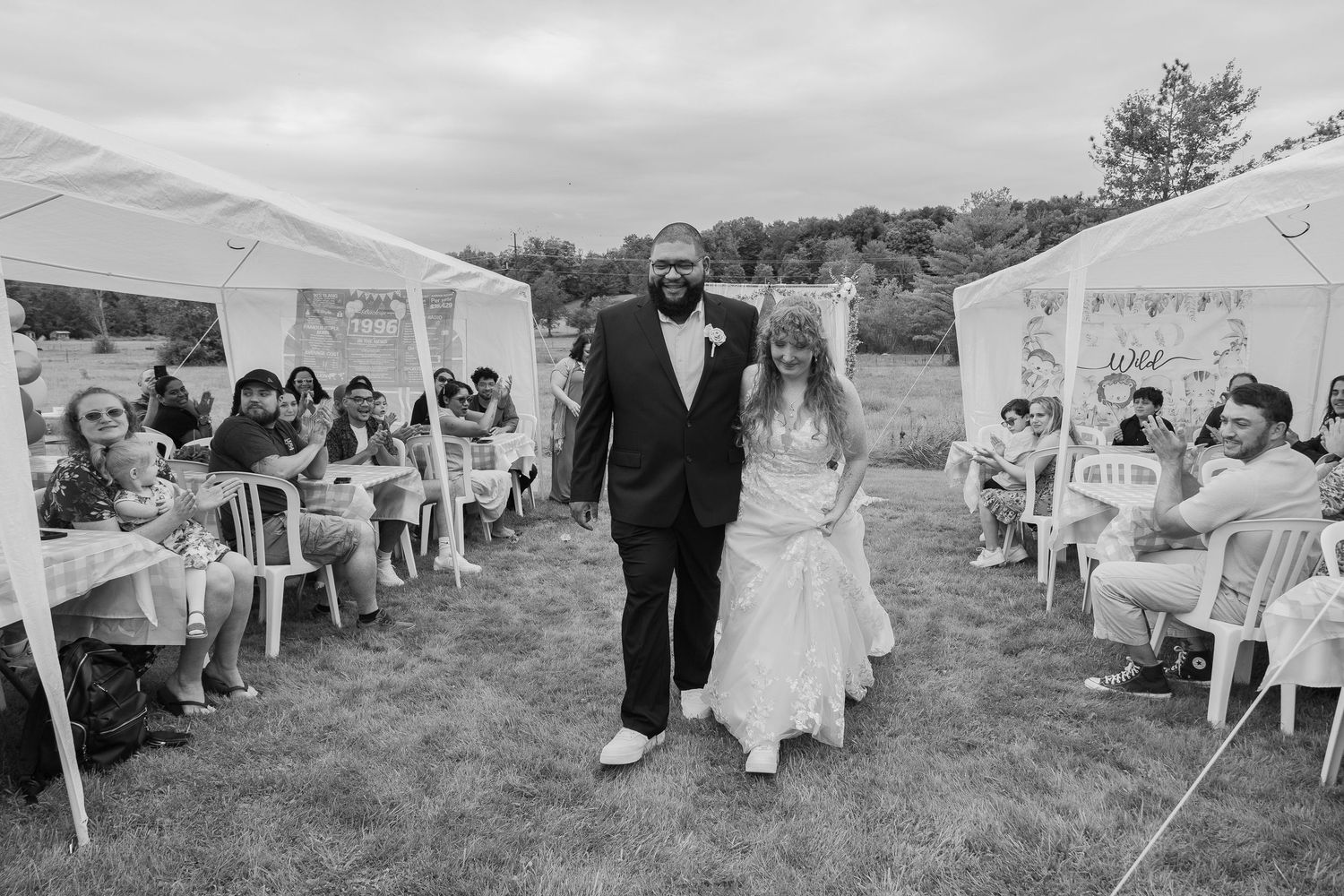 Black and white photo of a bride and groom walking together outdoors at a wedding reception with guests seated under tents.
