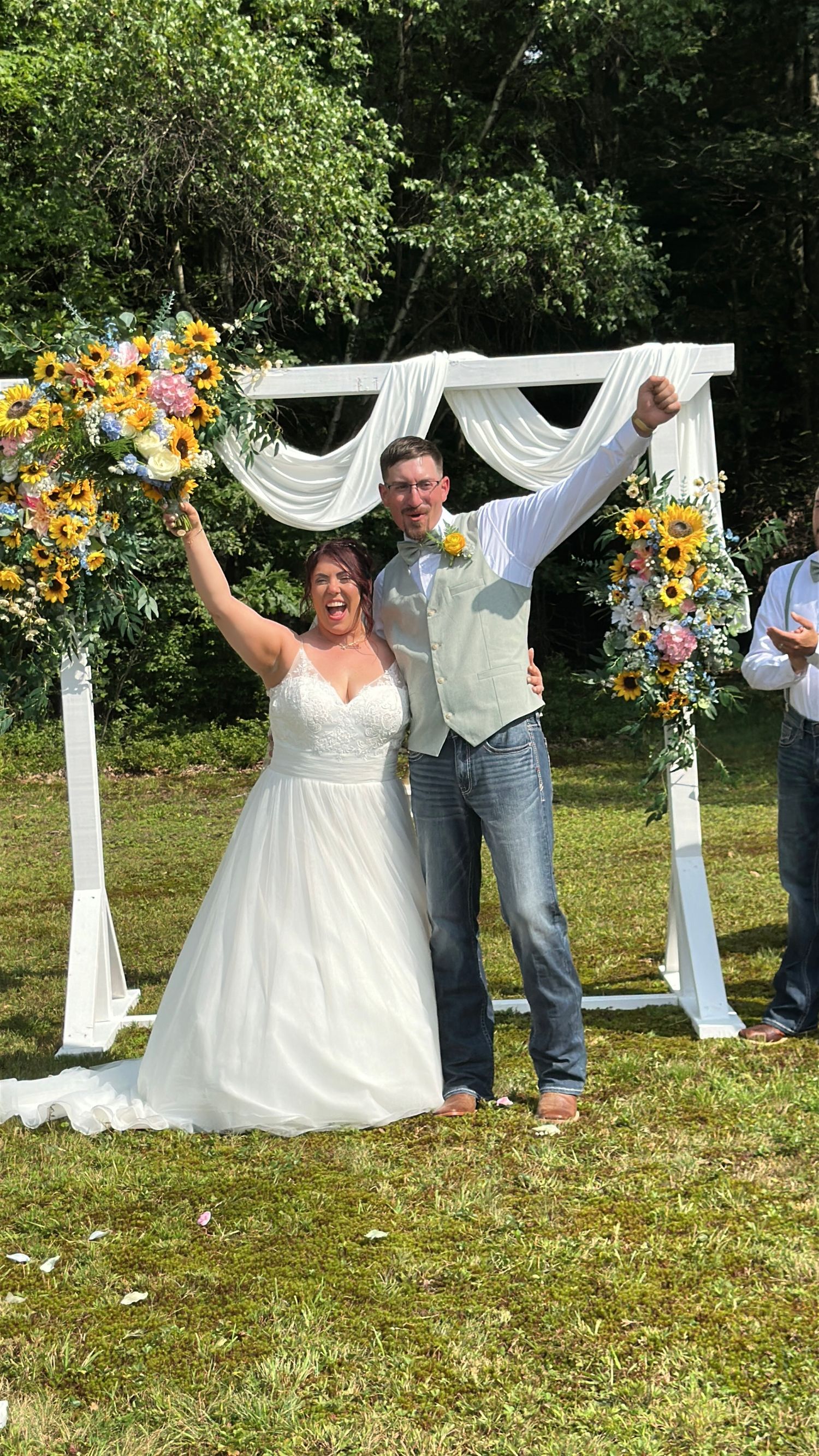 Joyful newlyweds celebrate under a white floral arch adorned with sunflowers, bride in white gown holding bouquet outdoors.