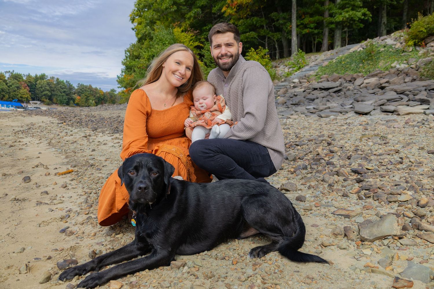 Family with baby and black Labrador posing on sandy beach with rocky shore and autumn trees in background.