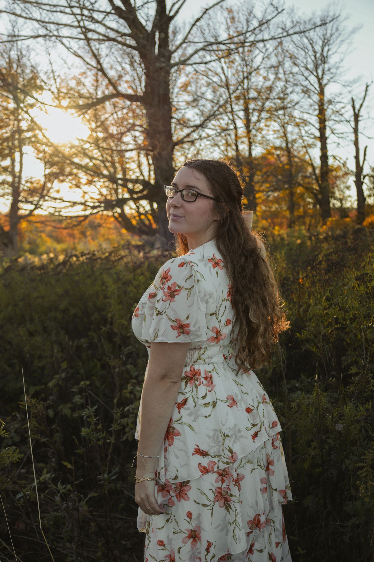 Young woman in floral dress with glasses poses outdoors at golden hour among autumn trees.