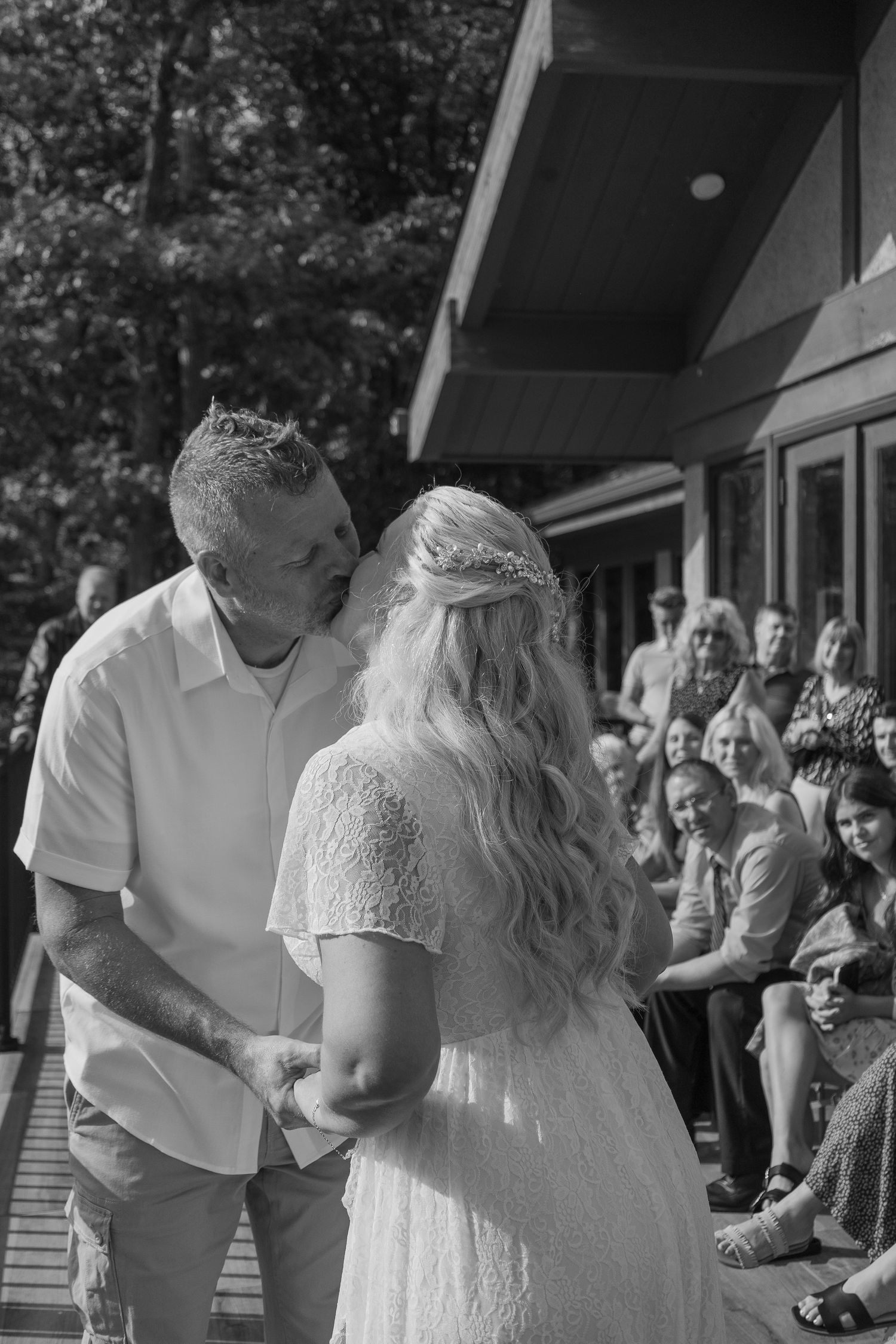 Black and white photo of bride and groom exchanging vows before smiling wedding guests outdoors.