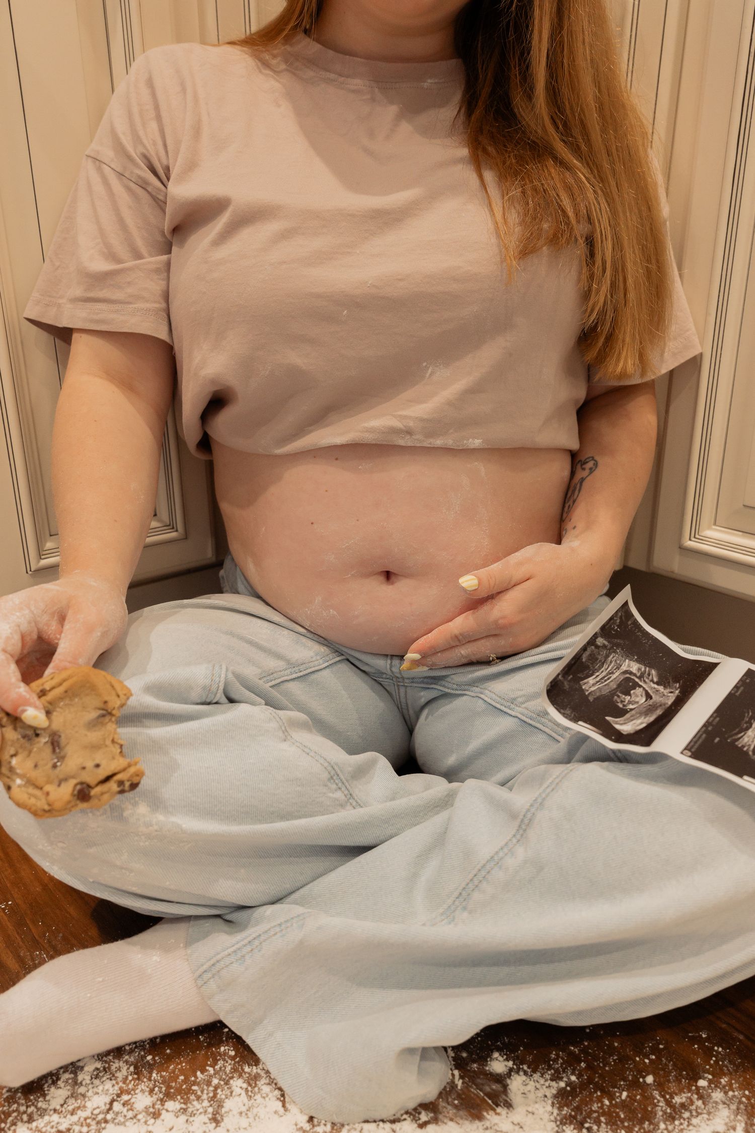 Pregnant woman sitting cross-legged holding a cookie and ultrasound photo, wearing crop top and jeans.