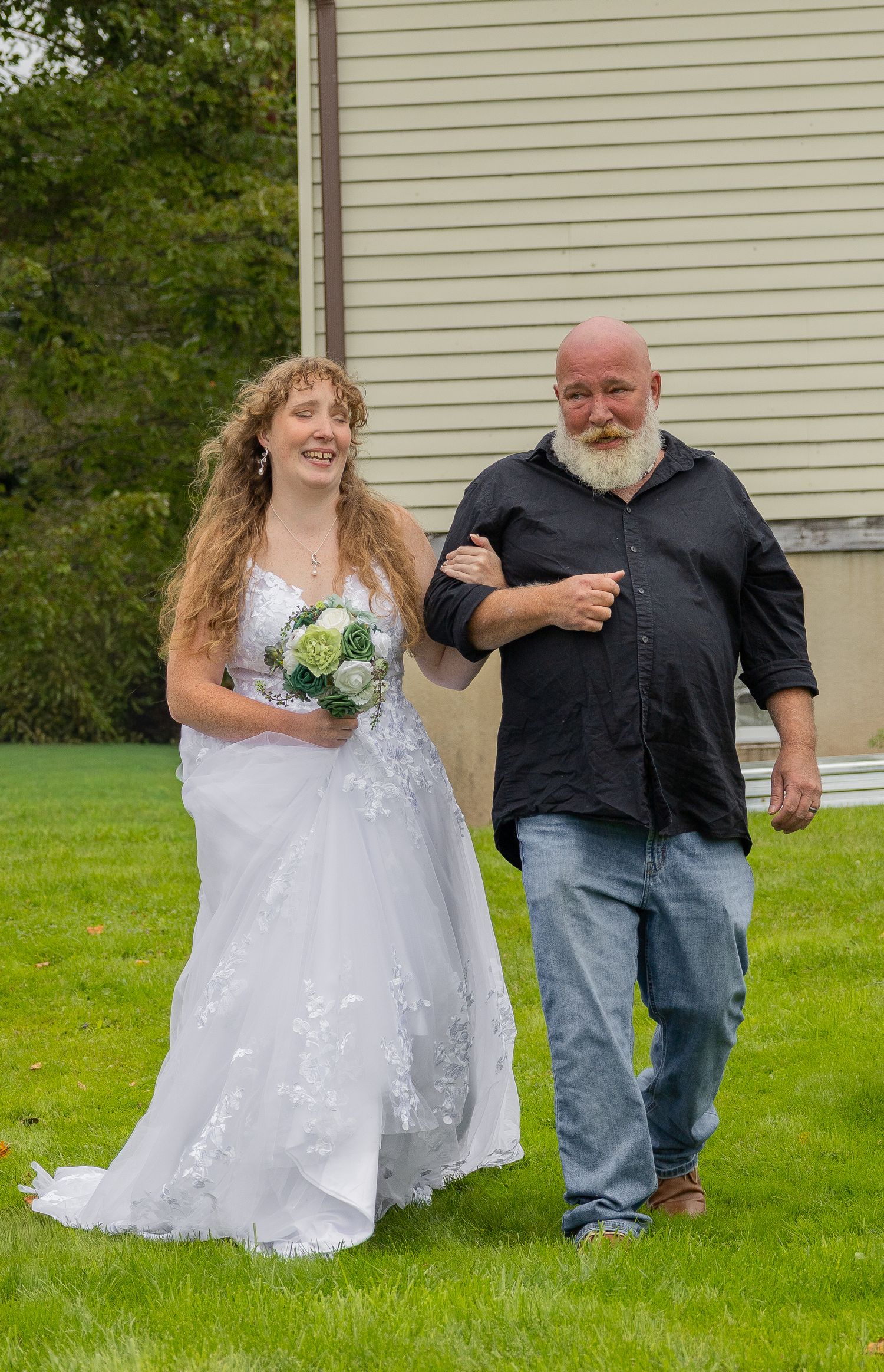 Smiling bride in white gown holding green bouquet walks arm-in-arm with bald bearded man in black shirt on green lawn.