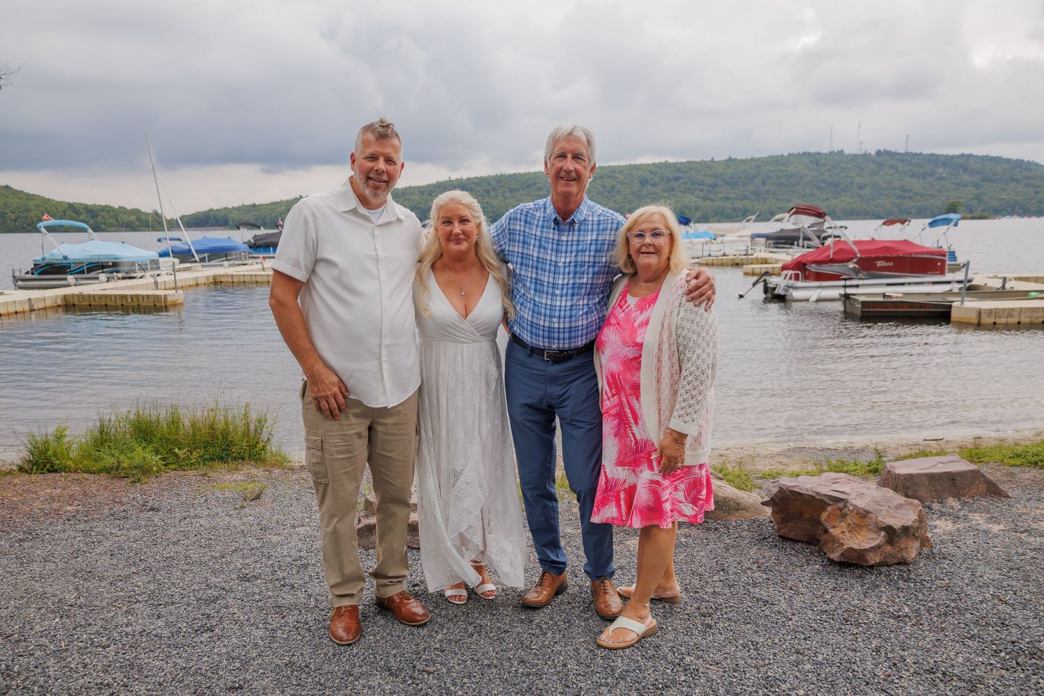 Two couples smiling together near a lakeside dock with boats and green hills in the background.