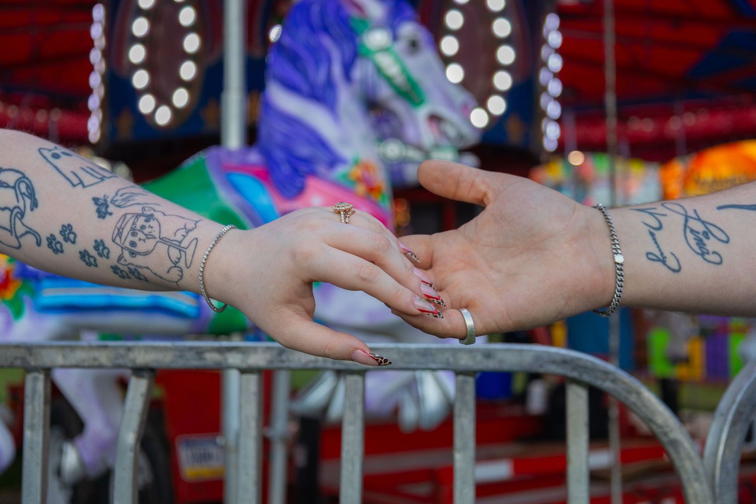 Two hands touching over a metal railing at a carnival, one with tattoos and a bracelet, with a carousel in the background.