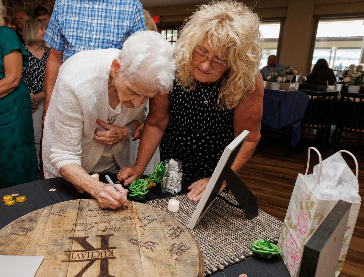 Two guests signing a wooden wedding guest book at a reception venue table.