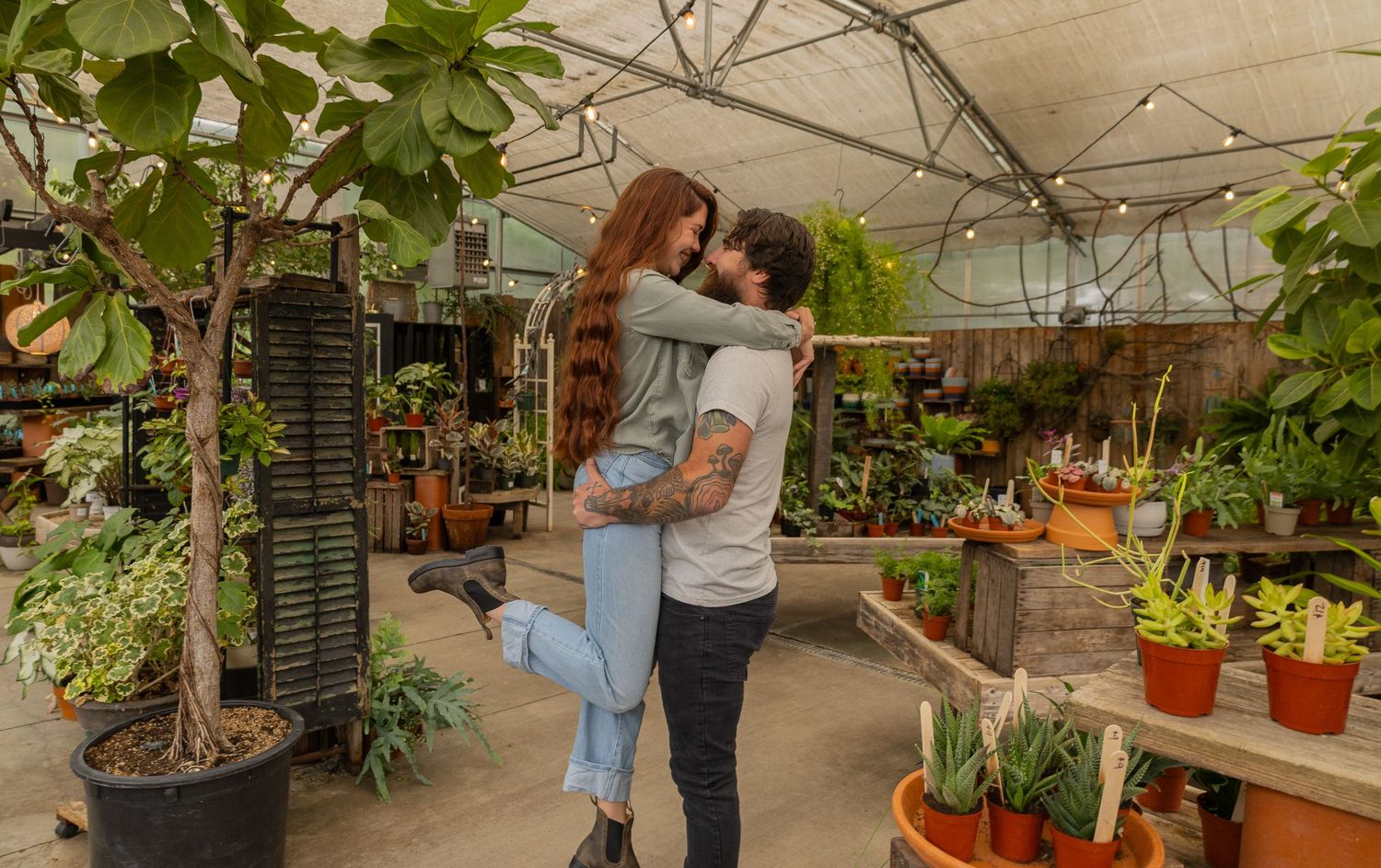 Couple embracing joyfully in a lush greenhouse garden center surrounded by potted plants and cacti.