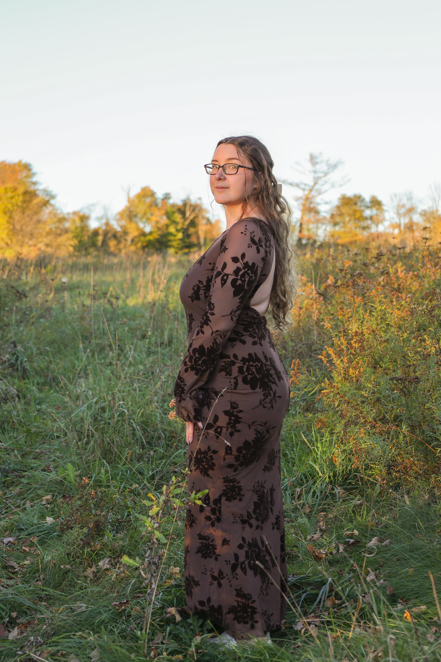 A young woman in a floral brown maxi dress and glasses stands in a lush green field during golden hour.