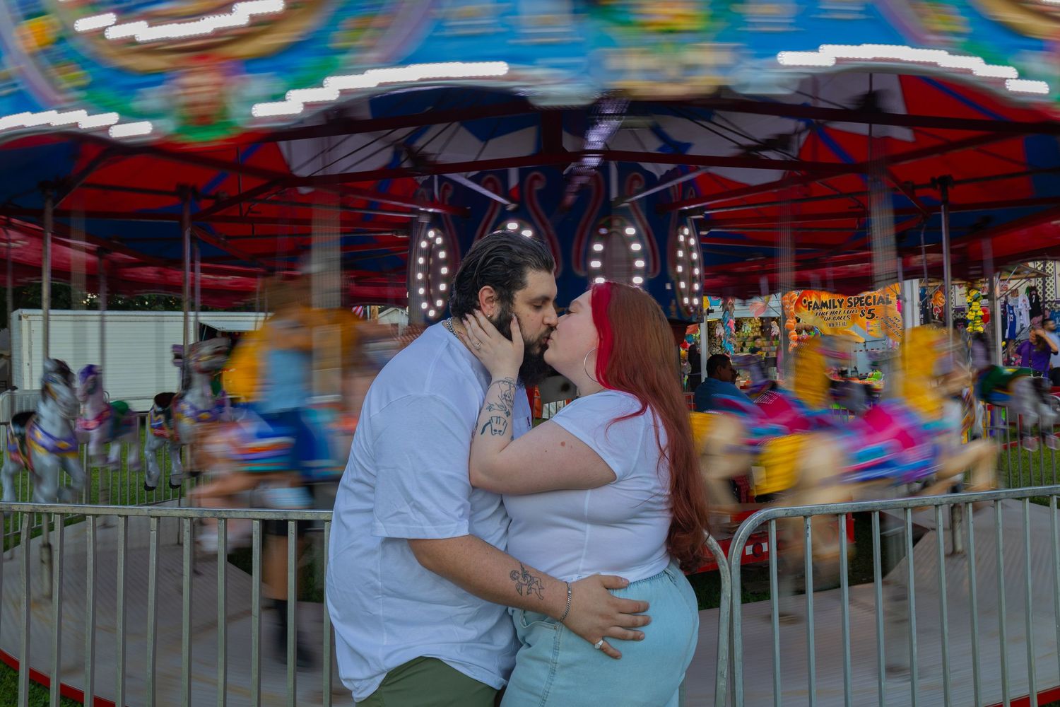Couple kissing in front of a spinning carousel at a colorful amusement park during dusk.