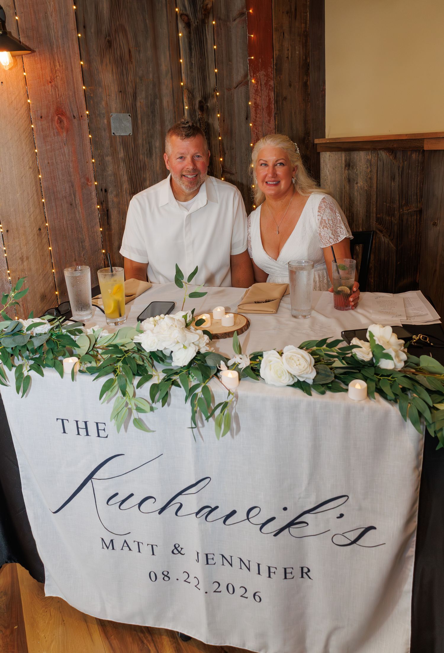 Matt and Jennifer Kuchavik sit smiling at floral sweetheart table at their 08-22-2026 wedding.