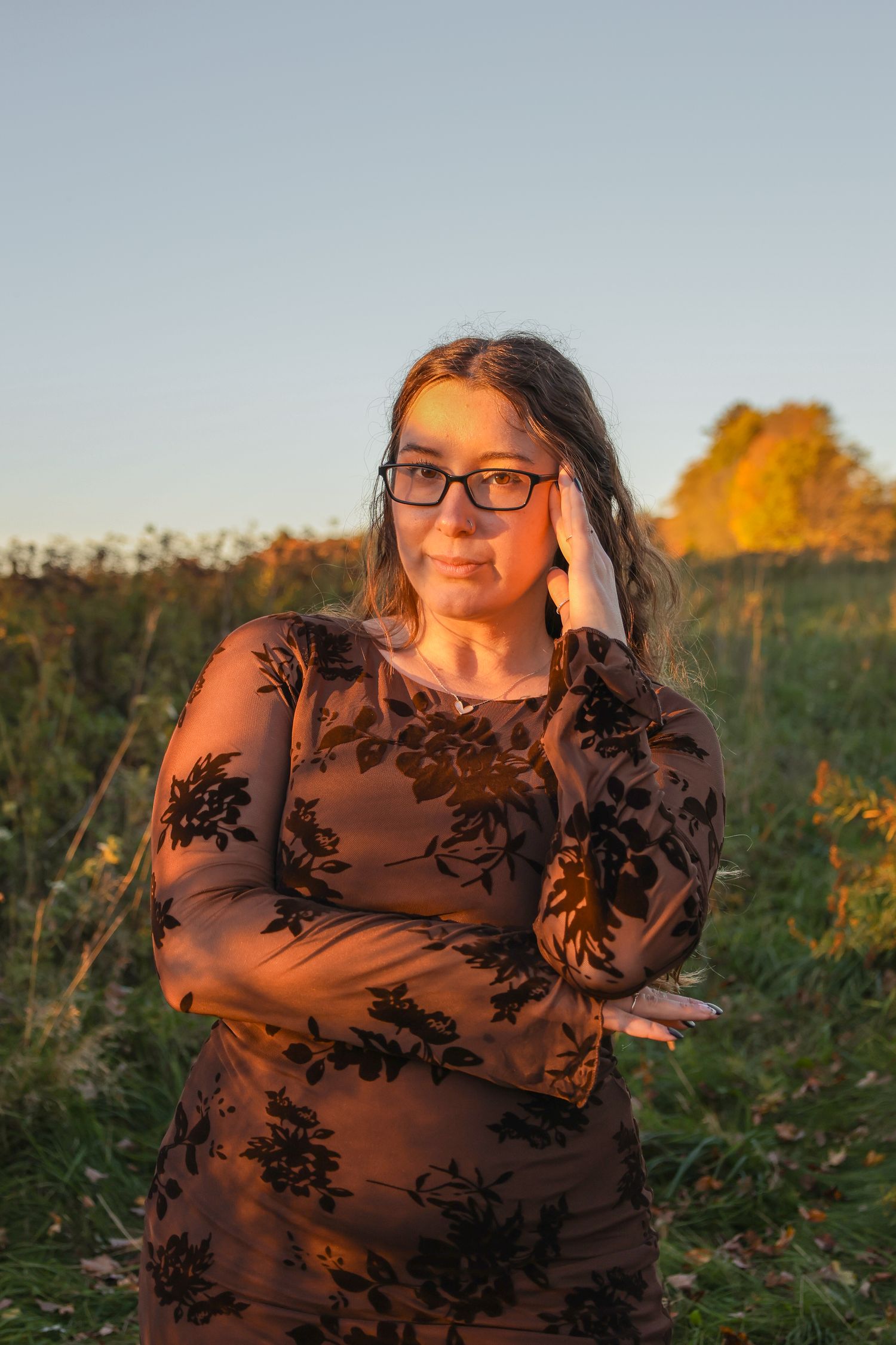 Woman in floral brown dress and glasses poses outdoors at golden hour with green field background.