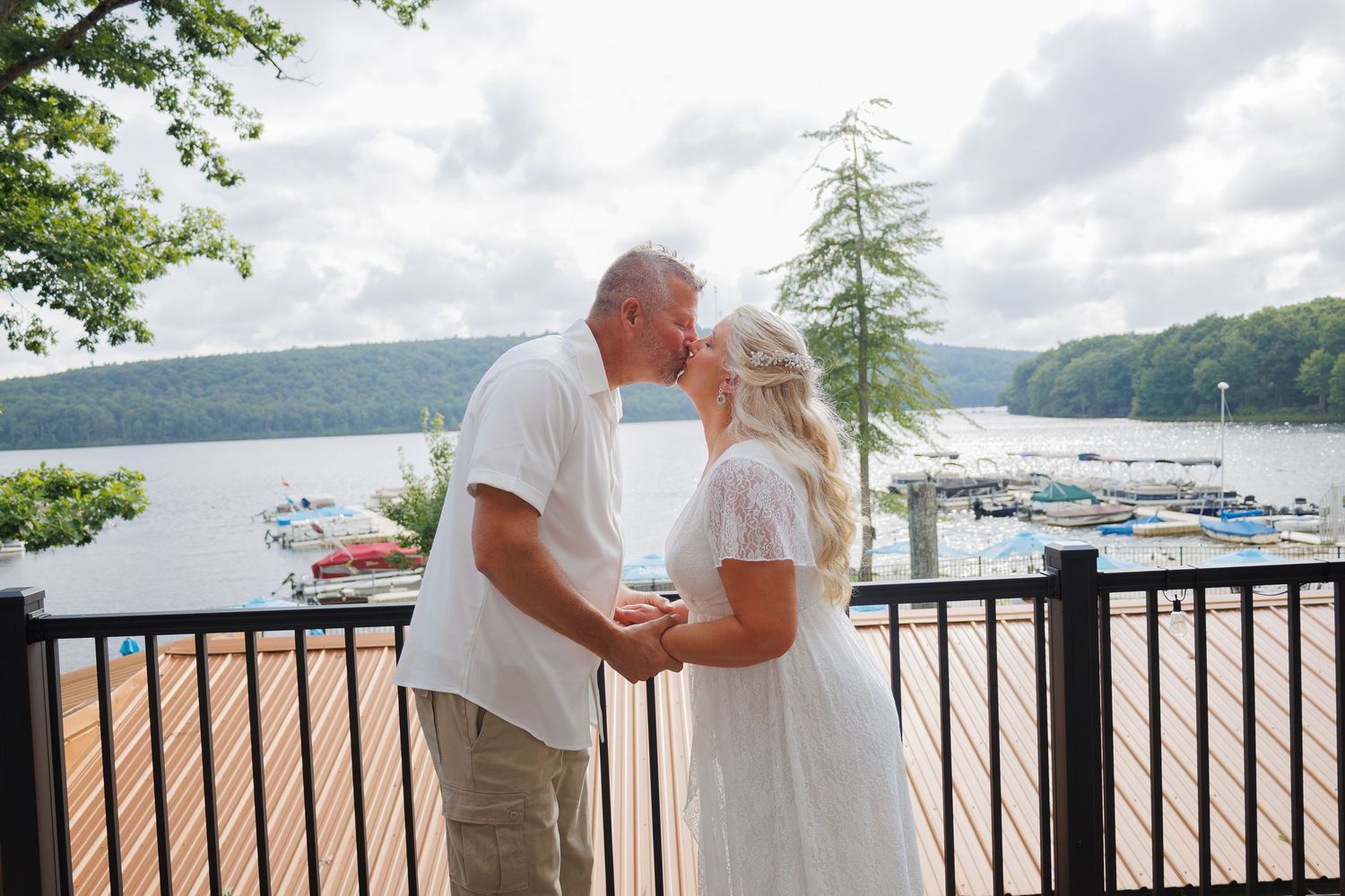 Couple sharing a romantic kiss on a lakeside deck with marina and lush green hills in the background.