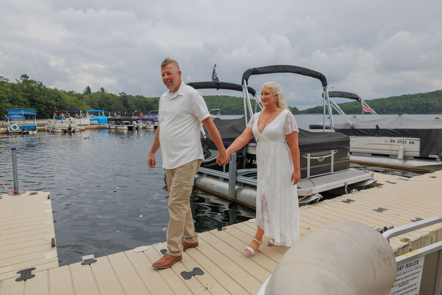 Couple holding hands on a lakeside dock with a pontoon boat and marina in the background on a cloudy day.