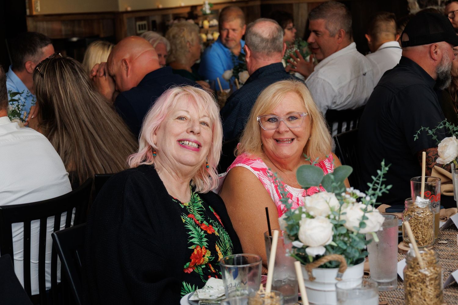 Two smiling women seated at a decorated banquet table during a festive indoor event or celebration.