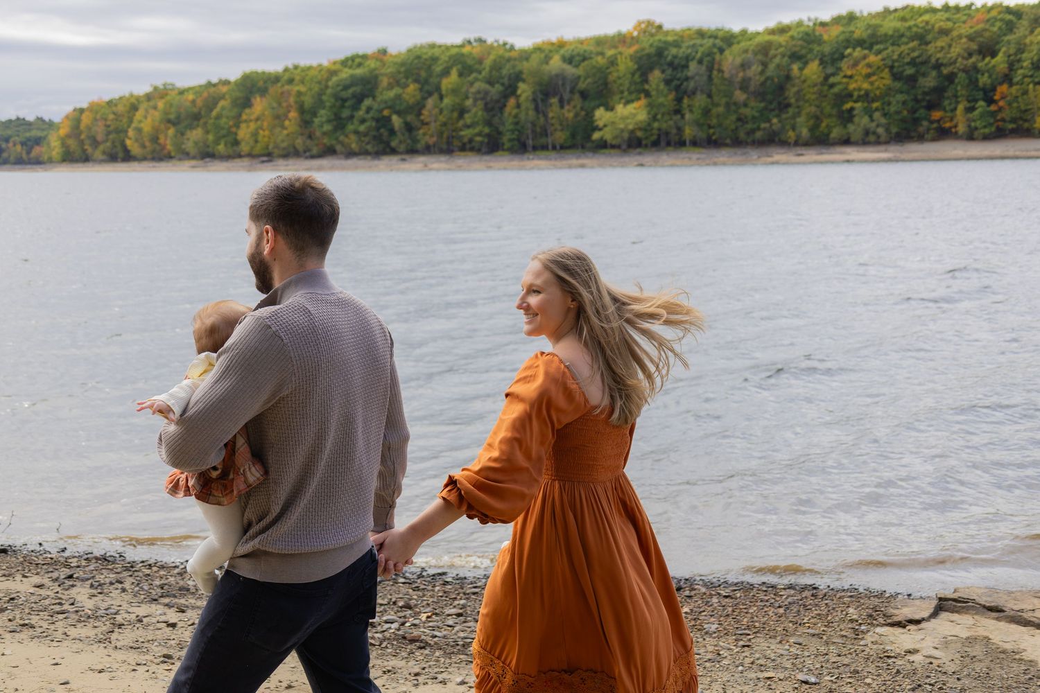 Family walking along a rocky riverbank in autumn, man holding baby, woman in orange dress, colorful fall trees in background.