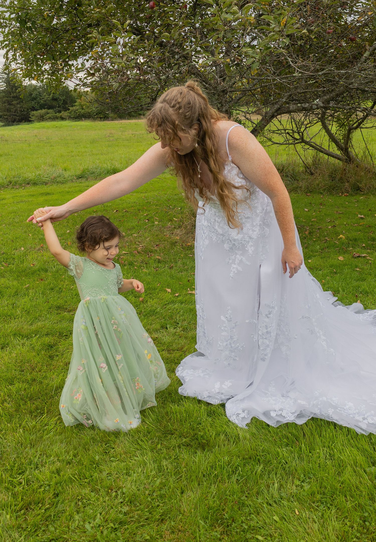 Bride in white lace gown twirling young flower girl in mint green dress on lush green lawn near tree.