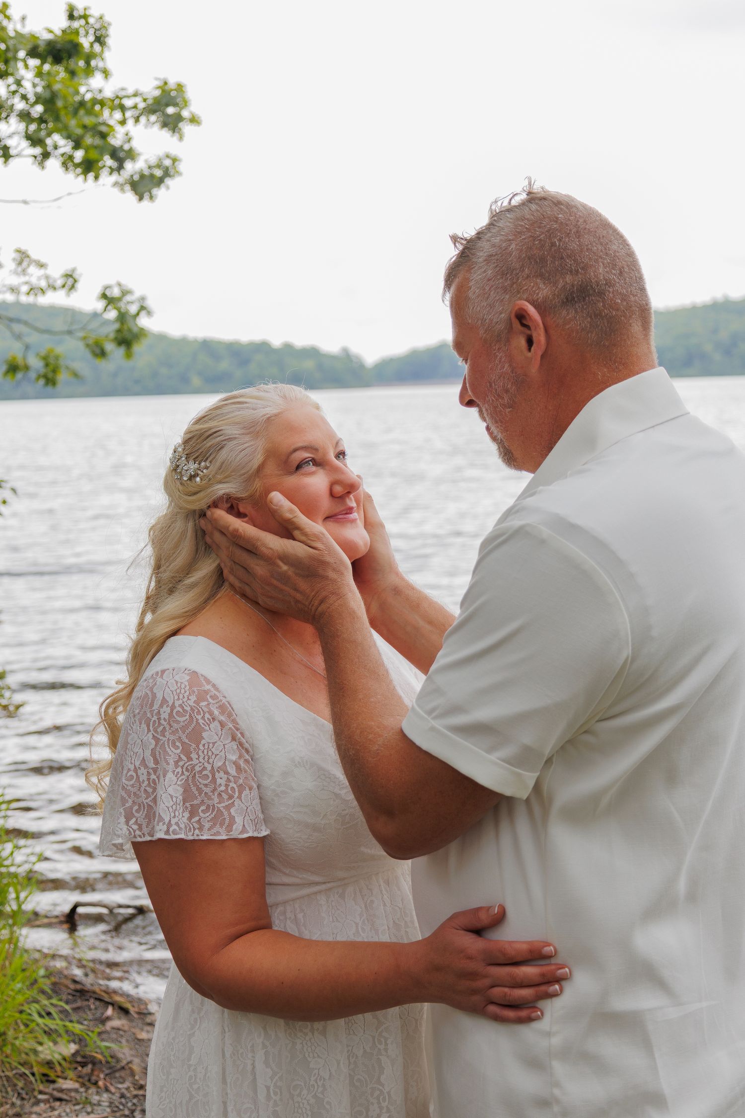 Groom tenderly holds brides face near a lakeside during an outdoor wedding ceremony.