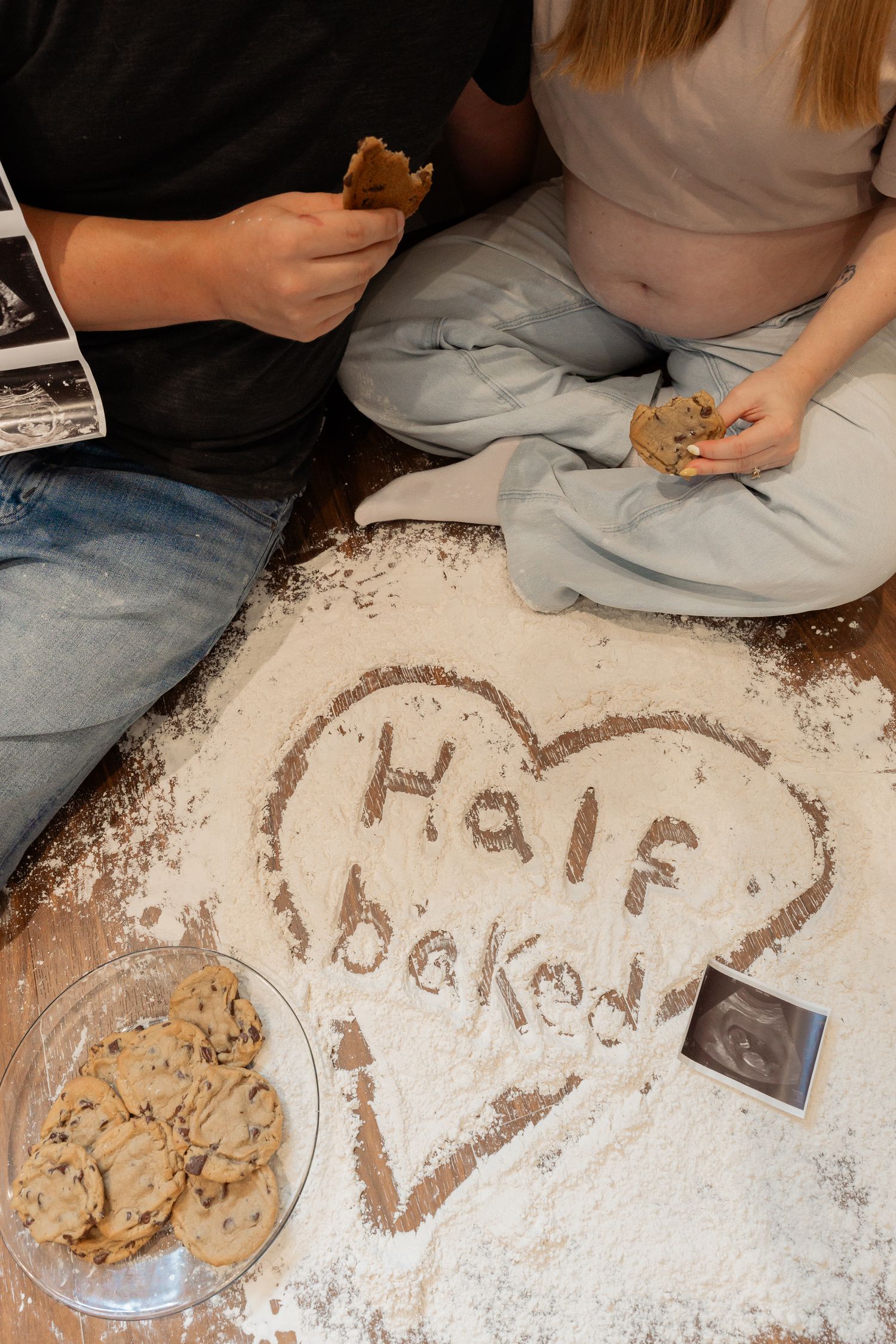 Couple sitting on floor with flour heart reading Half Baked, cookies, and ultrasound photos for pregnancy announcement.