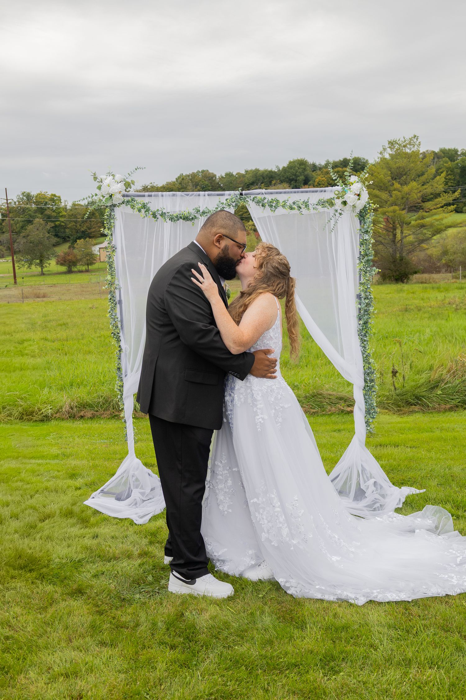 Bride and groom share first kiss under white floral arch on green lawn at outdoor wedding ceremony.