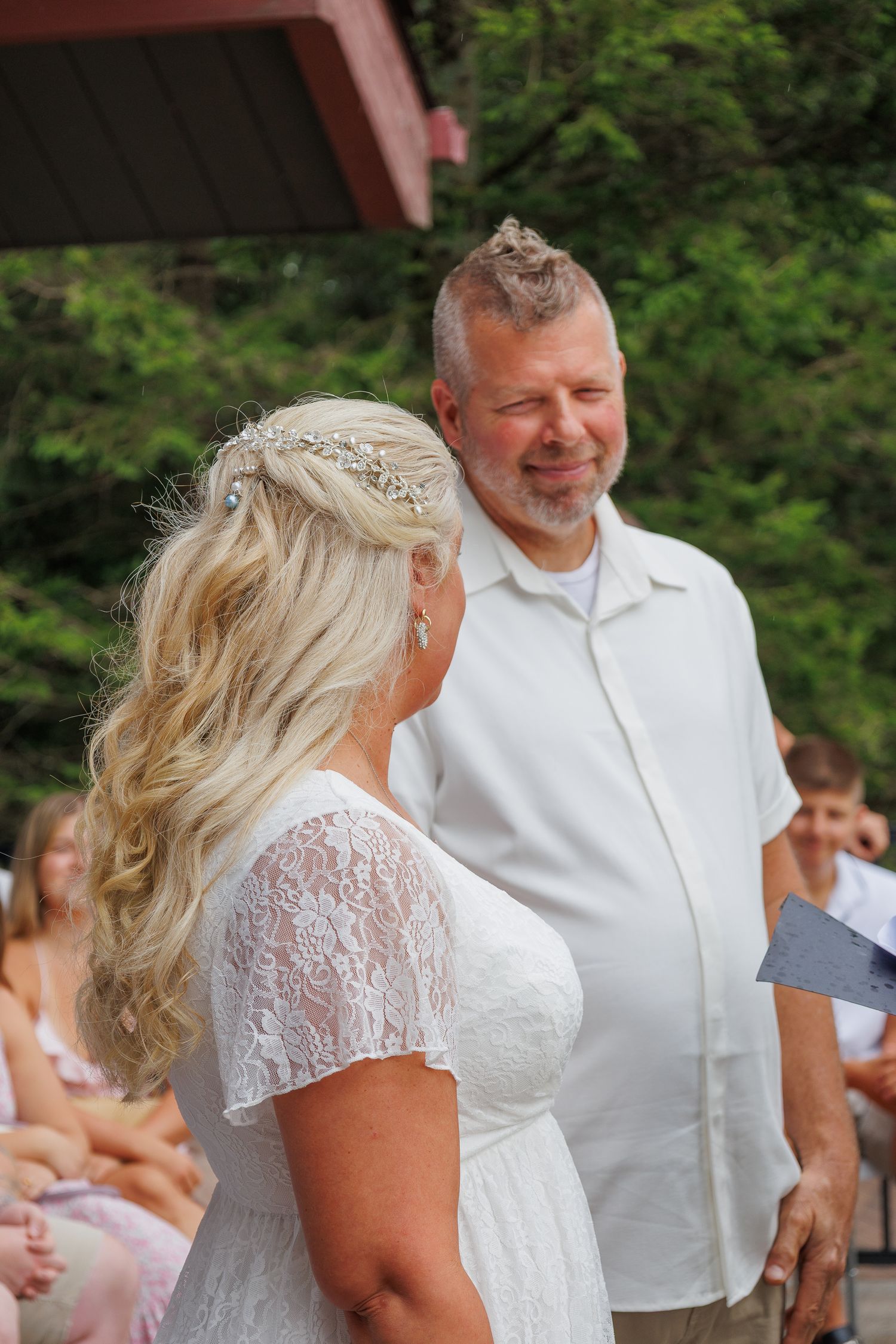 Groom smiles warmly at bride during outdoor wedding ceremony with guests seated in background.