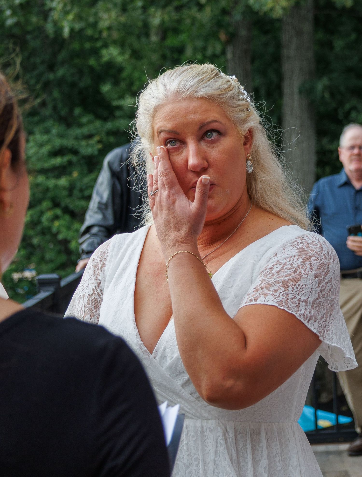 Emotional bride in white lace dress covers her mouth during outdoor wedding ceremony.