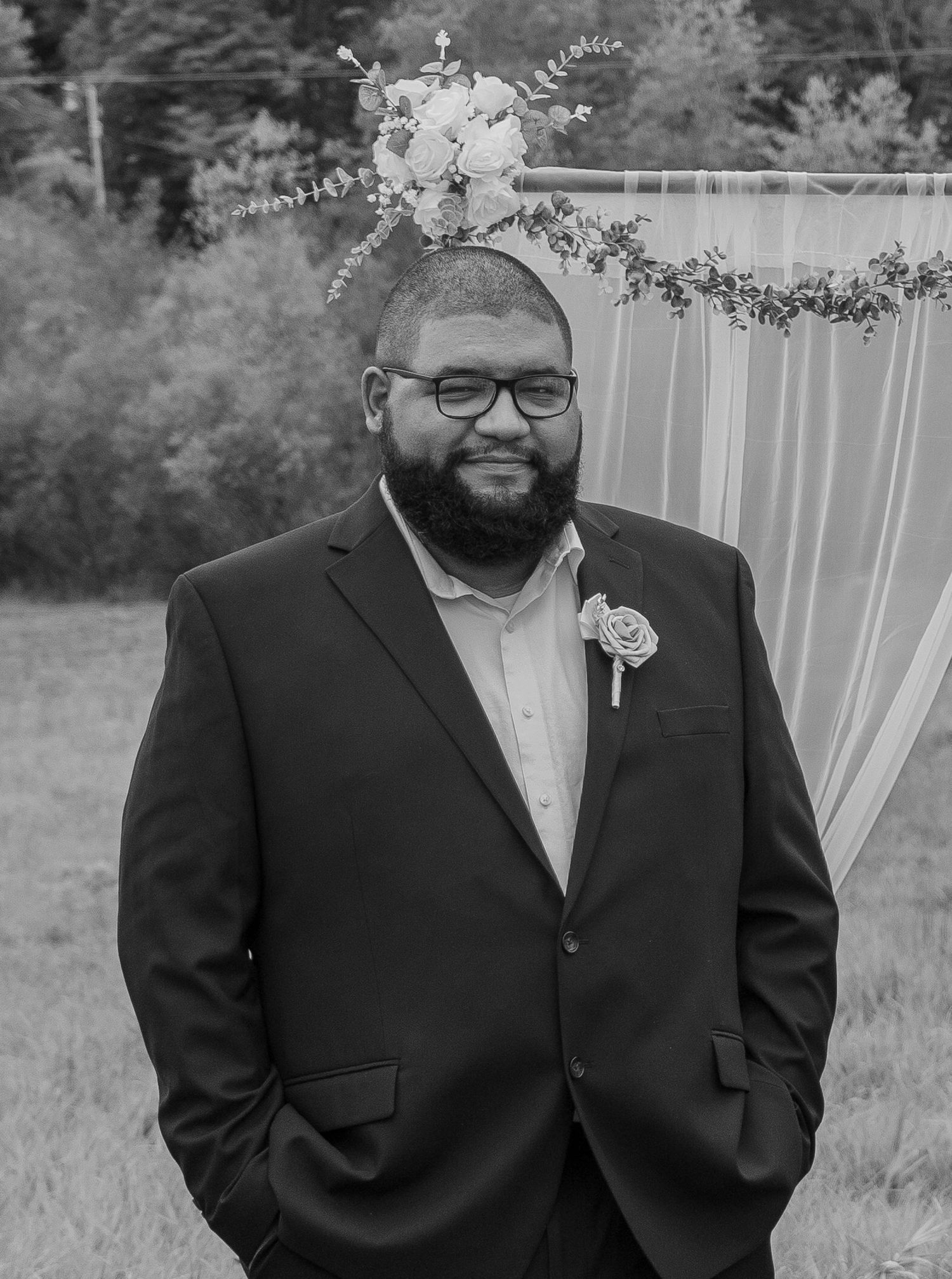 Man in suit with boutonniere smiling at outdoor wedding ceremony with floral arch backdrop.
