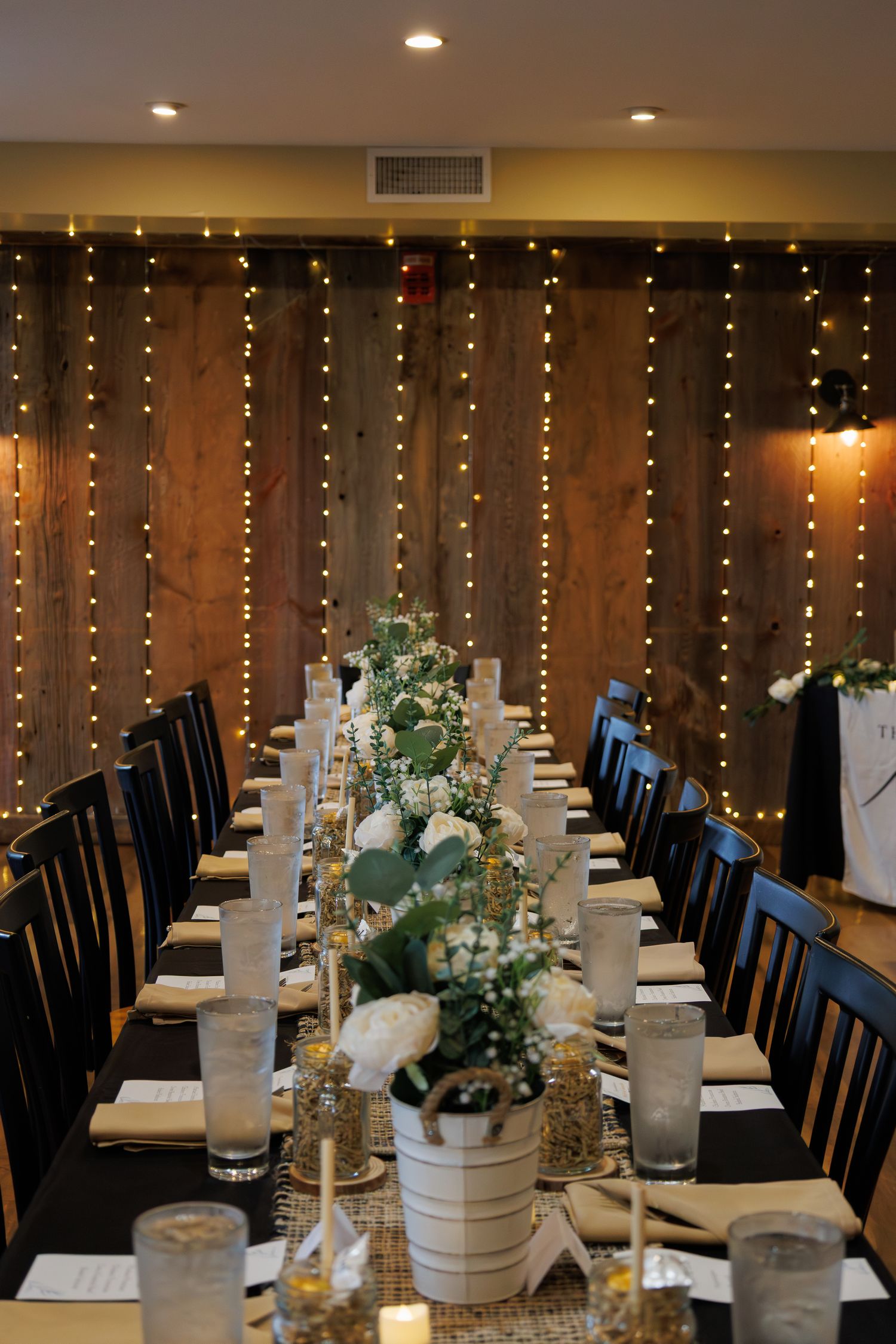 Long wedding reception table set with white floral centerpieces against a warm string light backdrop.