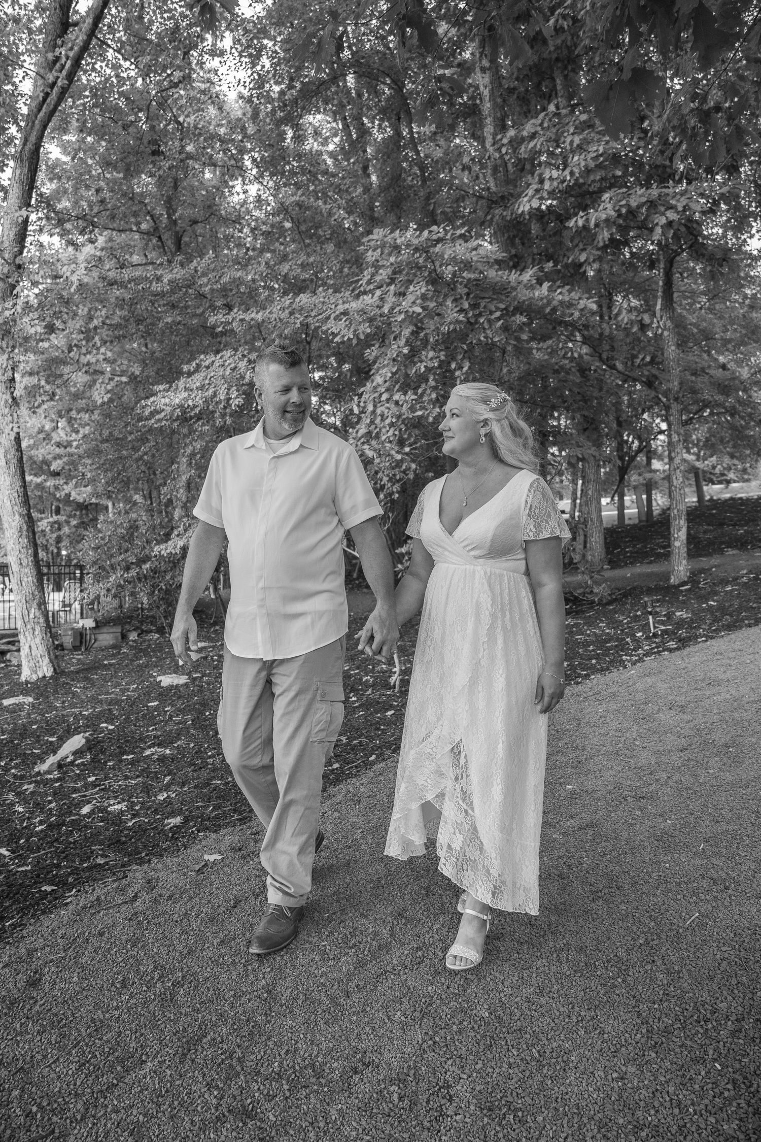 Bride and groom walk hand in hand along a tree-lined path in black and white wedding photo.