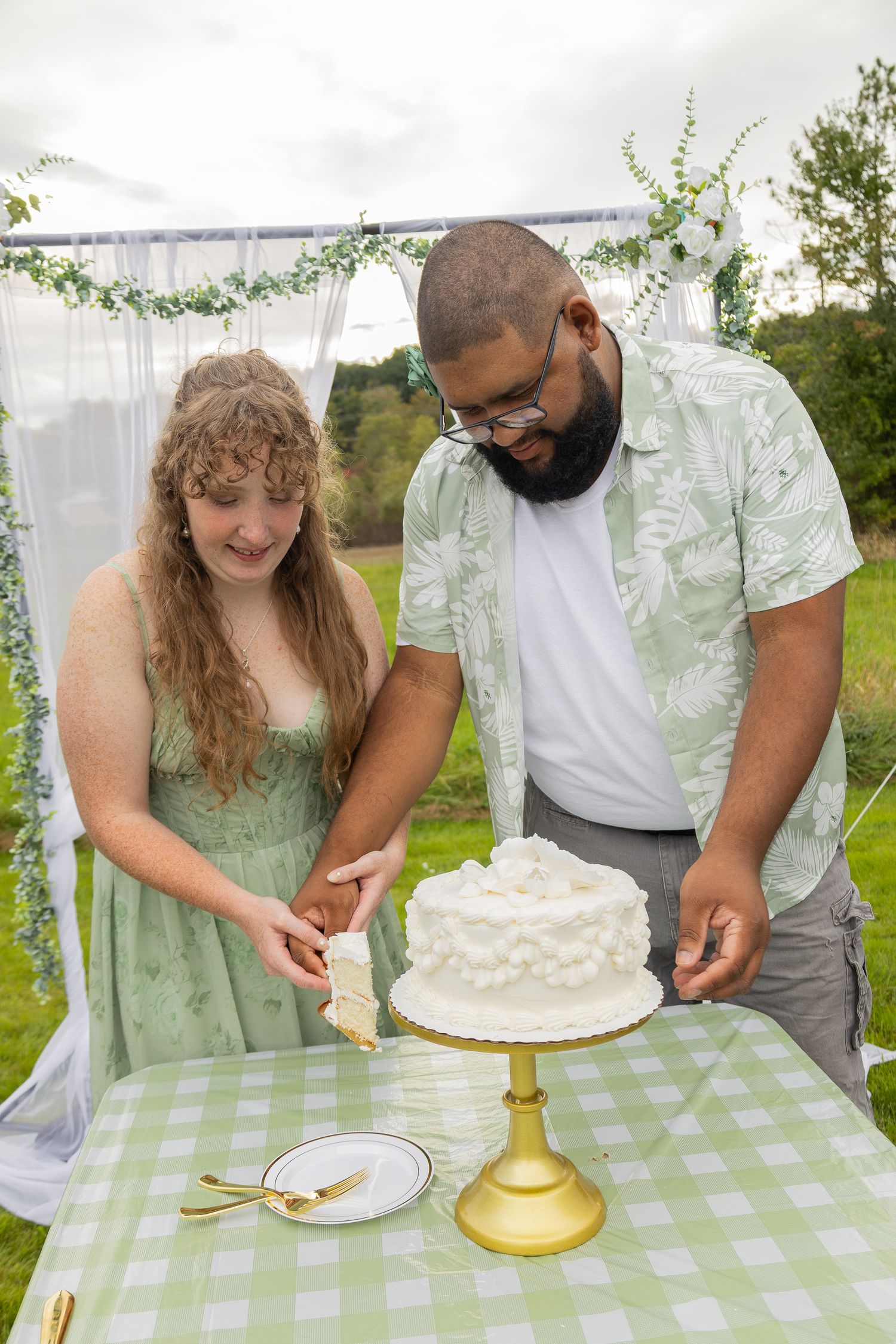 Couple cutting white wedding cake outdoors at green gingham table under floral arch.