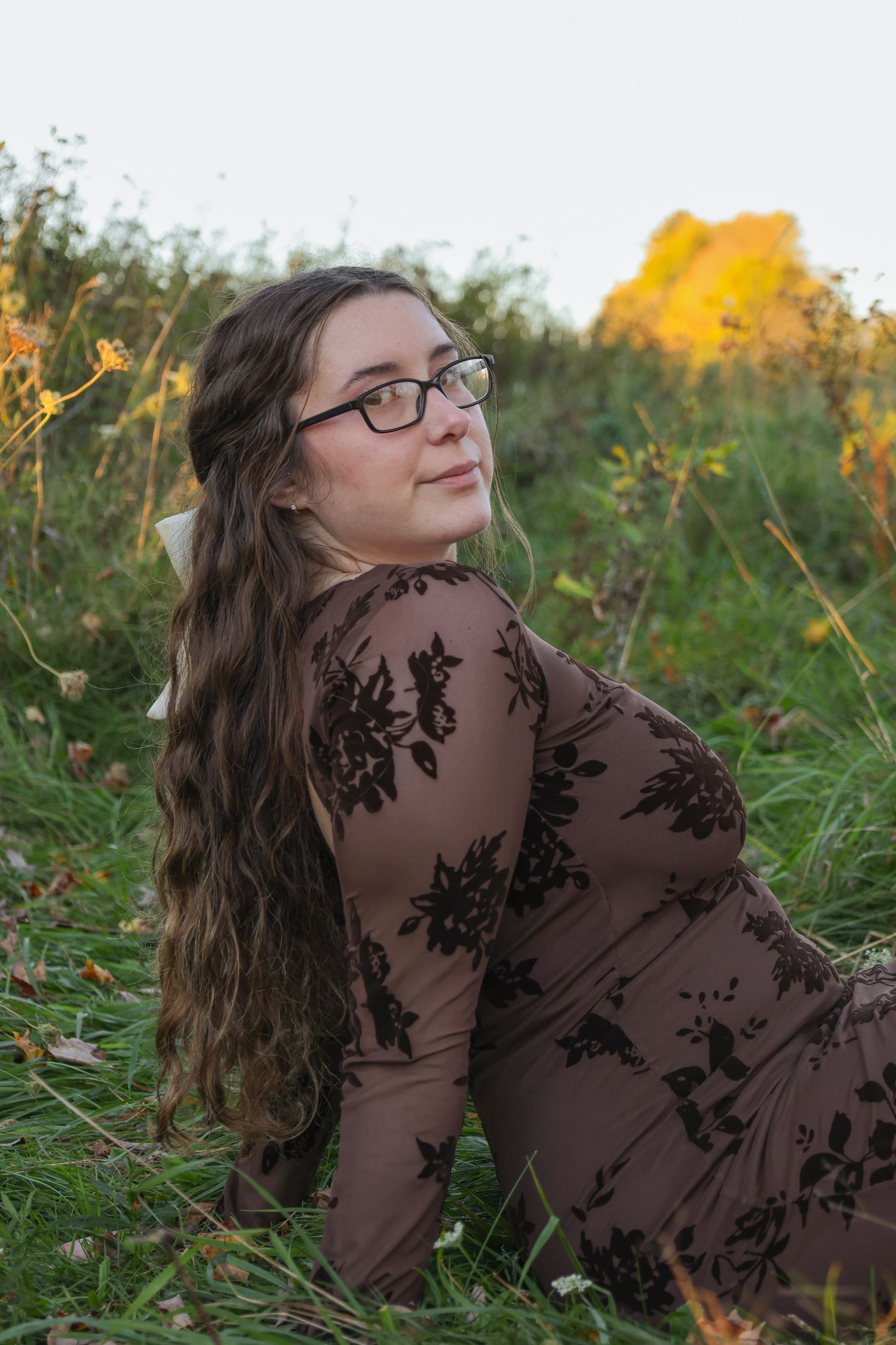 Young woman with long hair and glasses in a floral brown dress sitting in a green field with autumn foliage.