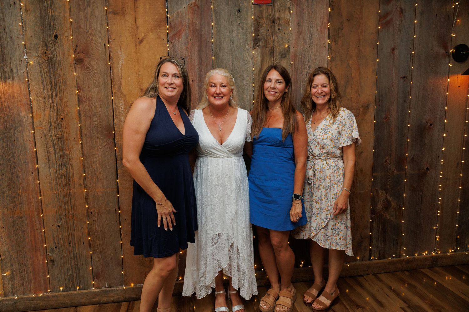 Four women smiling together in front of a rustic wood wall with string lights at an indoor event.