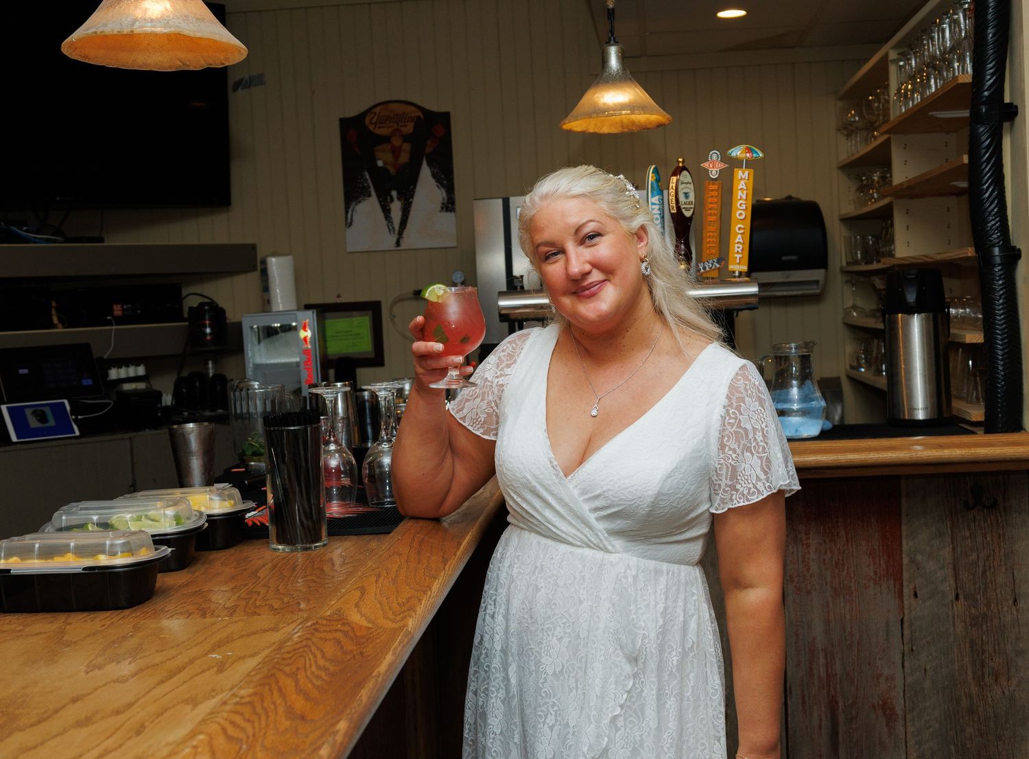 Bride in white dress smiles while raising a cocktail at a rustic wedding reception bar.