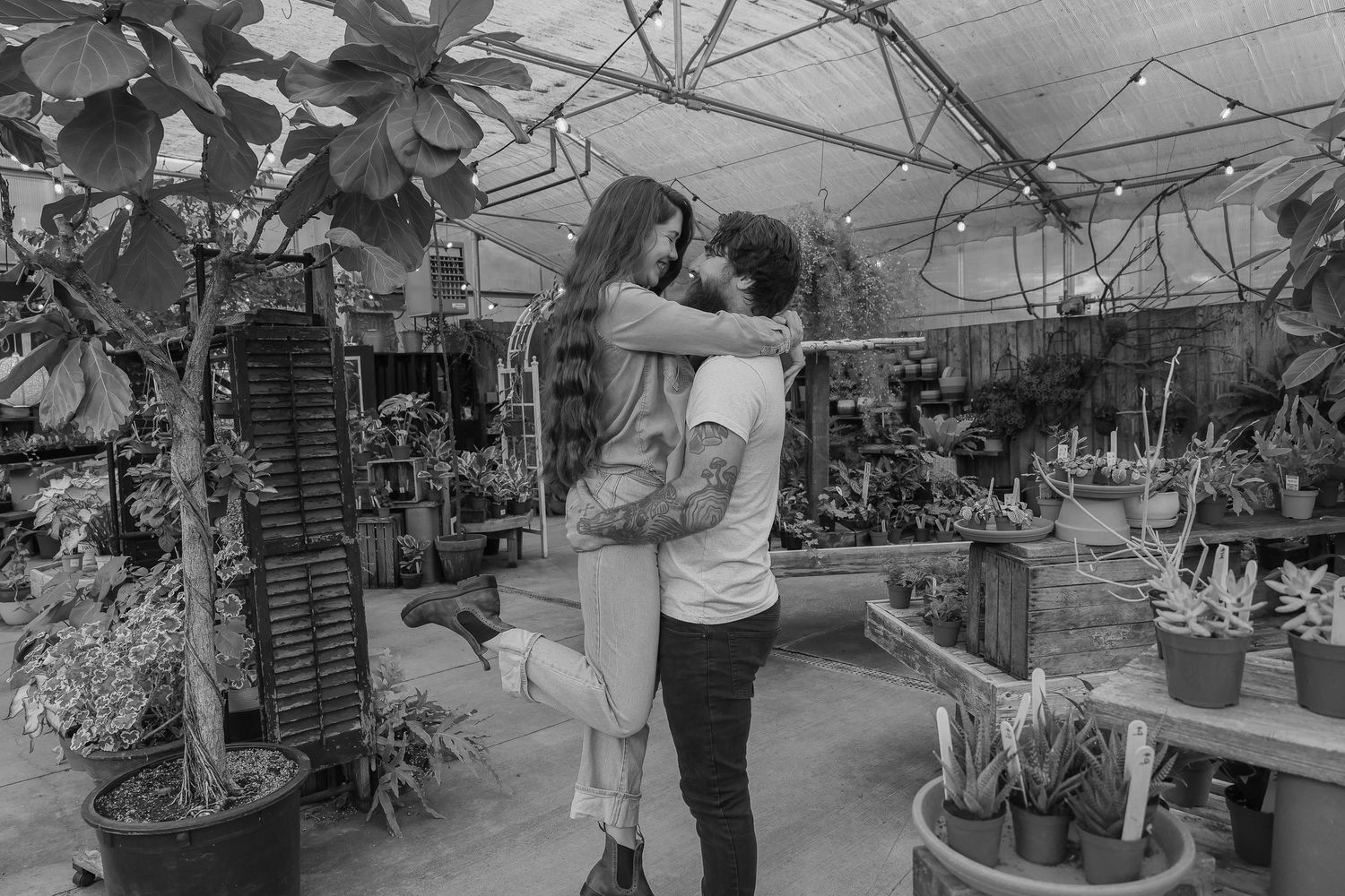 Couple embracing and lifting each other in a lush greenhouse surrounded by tropical plants, black and white photo.