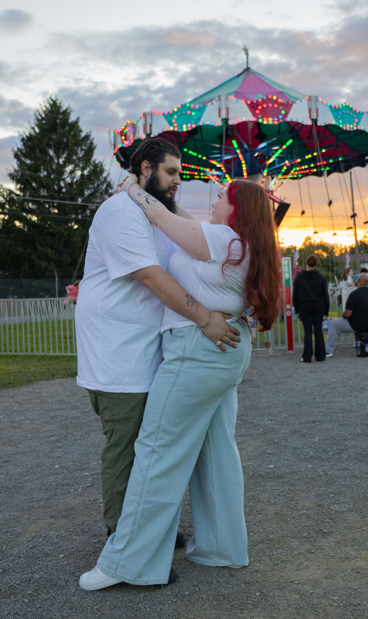 Couple embracing romantically at a carnival at sunset, with colorful carousel lights glowing behind them.