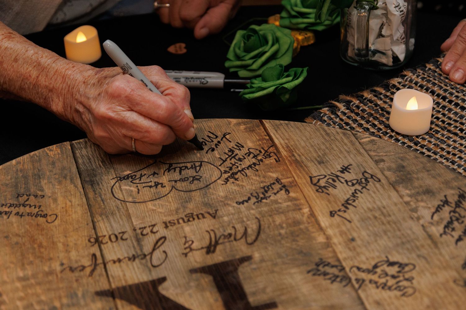 Guests signing a wooden barrel lid with a marker at a wedding reception, with candles and floral decor nearby.
