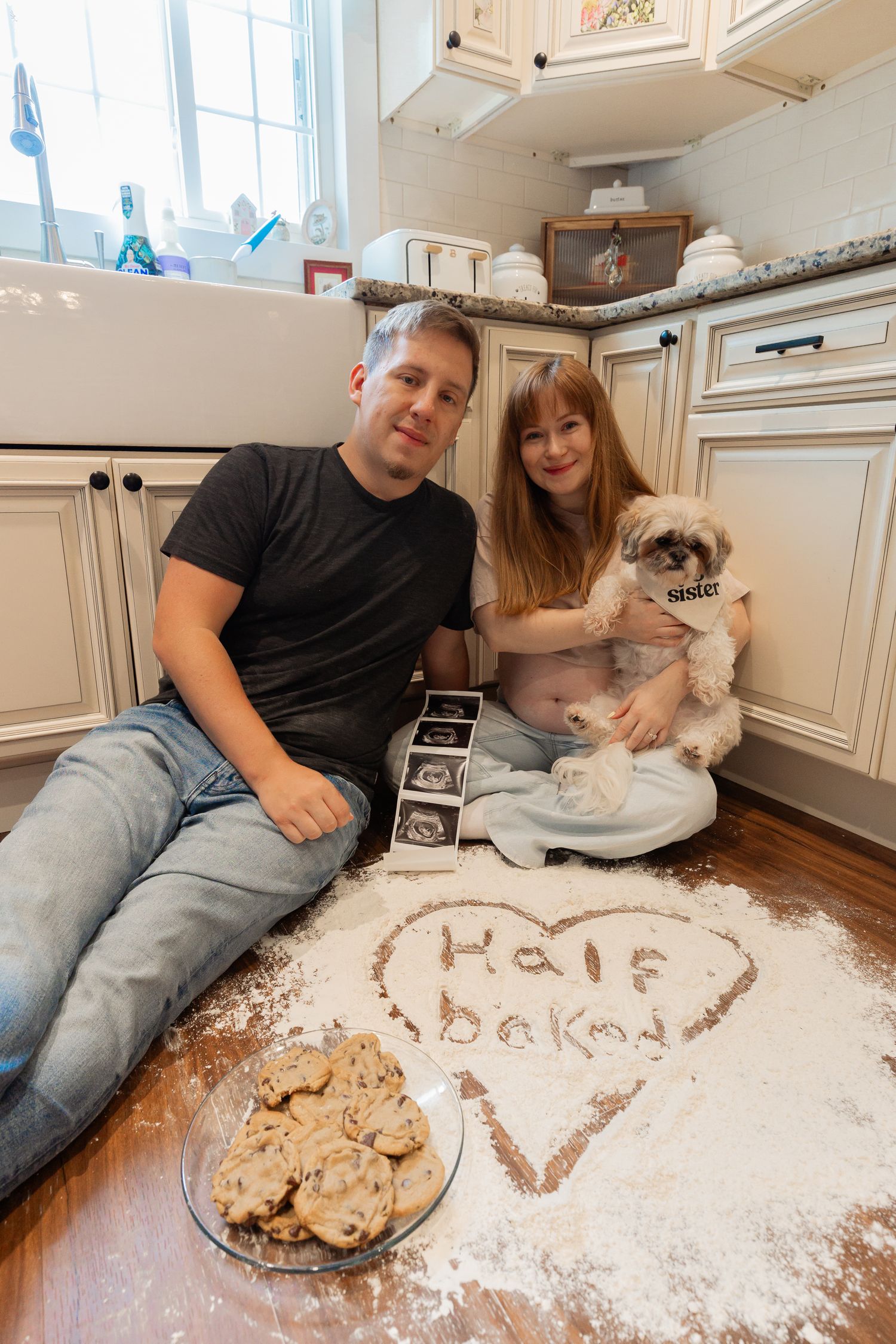 Couple sits on kitchen floor with dog and flour heart reading Half Baked for a creative pregnancy announcement photo.