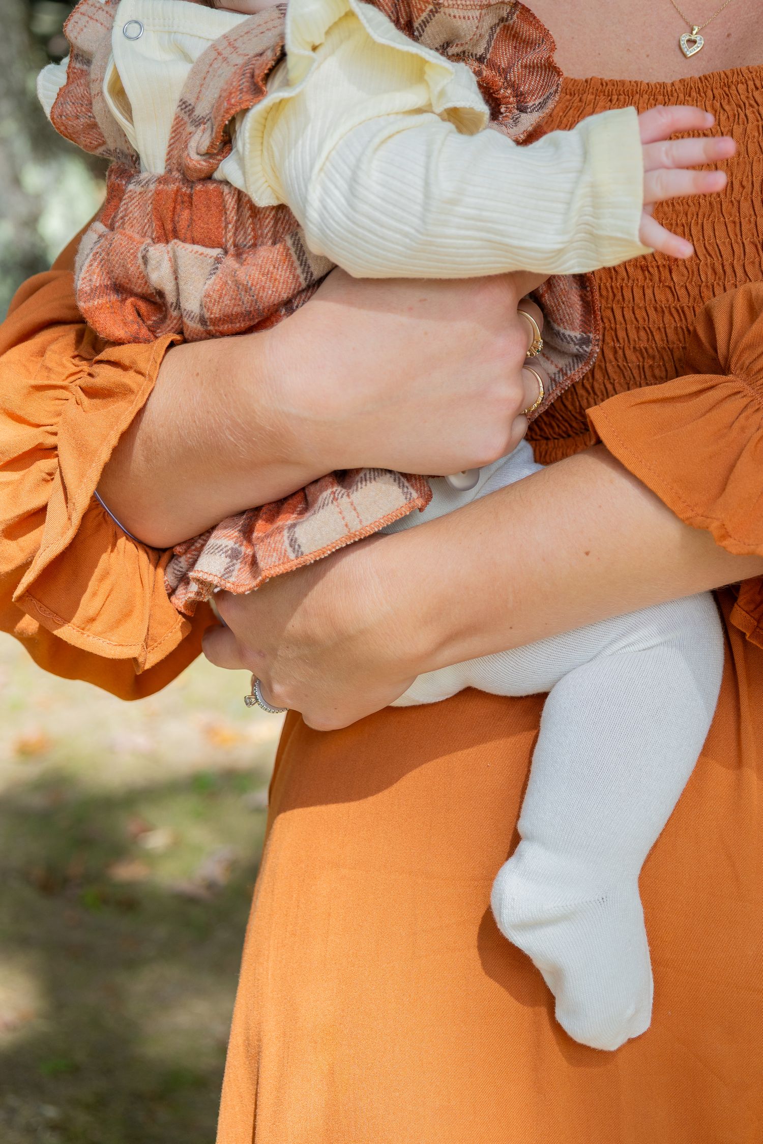 A mother in an orange dress cradles her newborn baby wrapped in a plaid blanket outdoors.