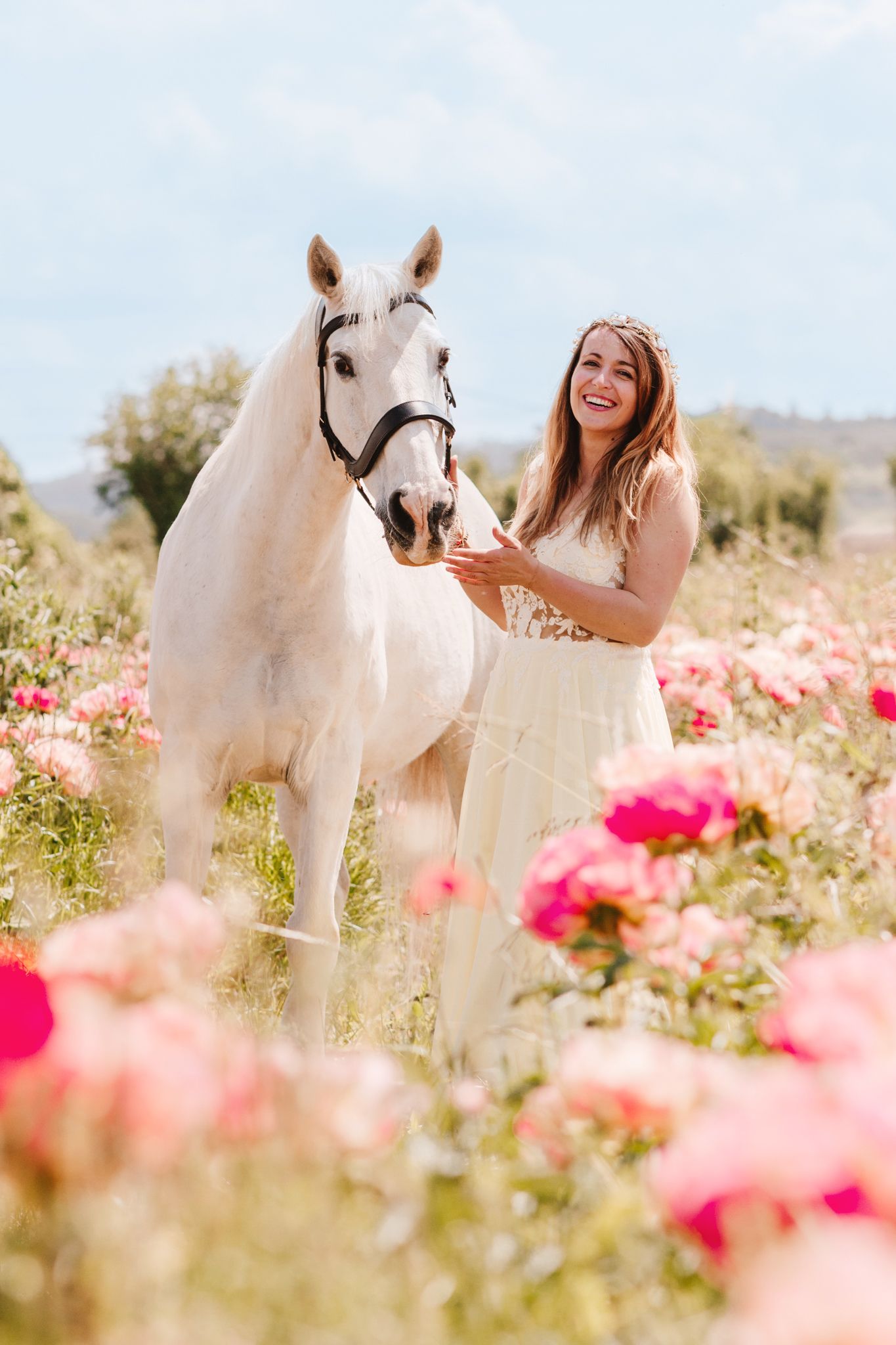Smiling woman in white dress feeding white horse in vibrant pink flower field.