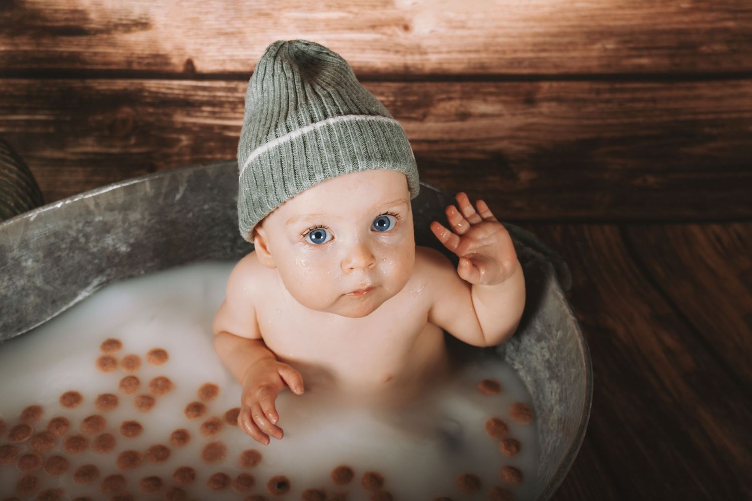 Adorable blue-eyed baby in gray knit hat sitting in vintage milk bath with rose petals.