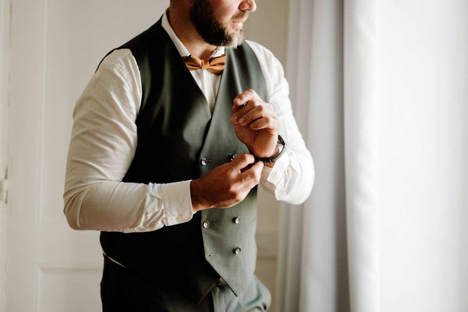 Groom in dark green vest and brown bow tie adjusting cufflinks while getting ready for wedding.