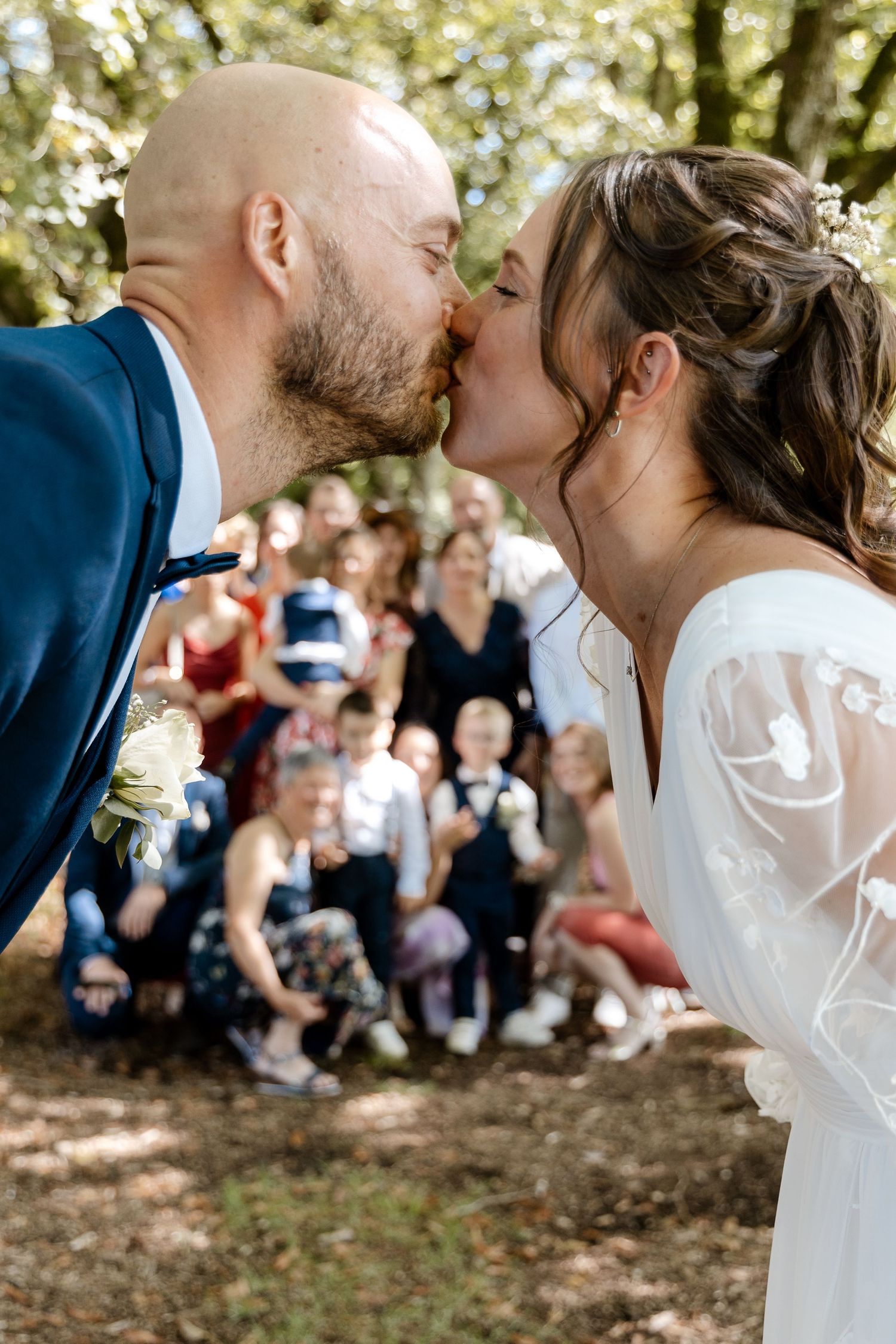 Bride and bald groom sharing a kiss with wedding guests gathered in background outdoors.