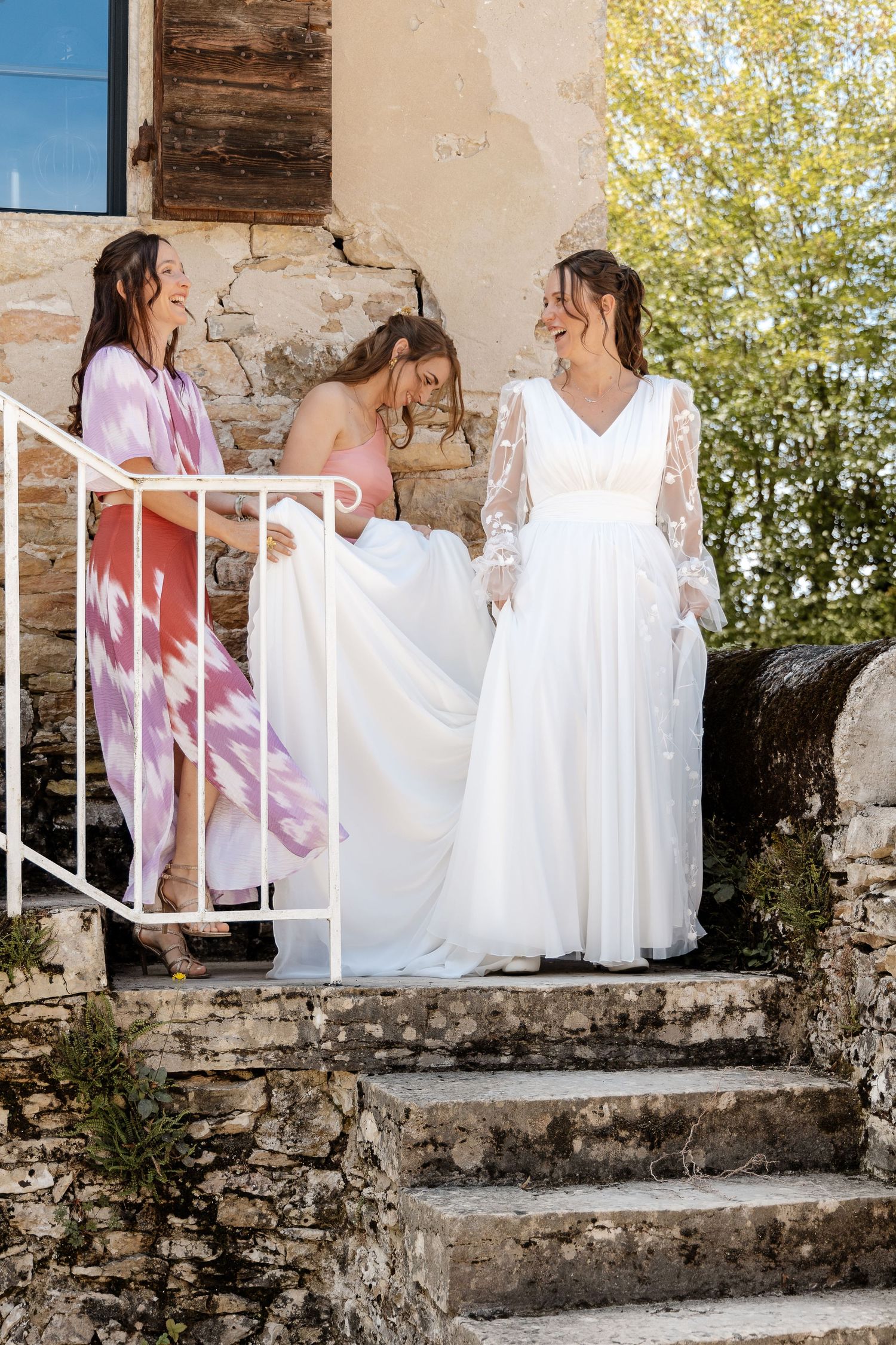 Bride in white lace gown with bridesmaids on rustic stone steps at outdoor wedding venue.