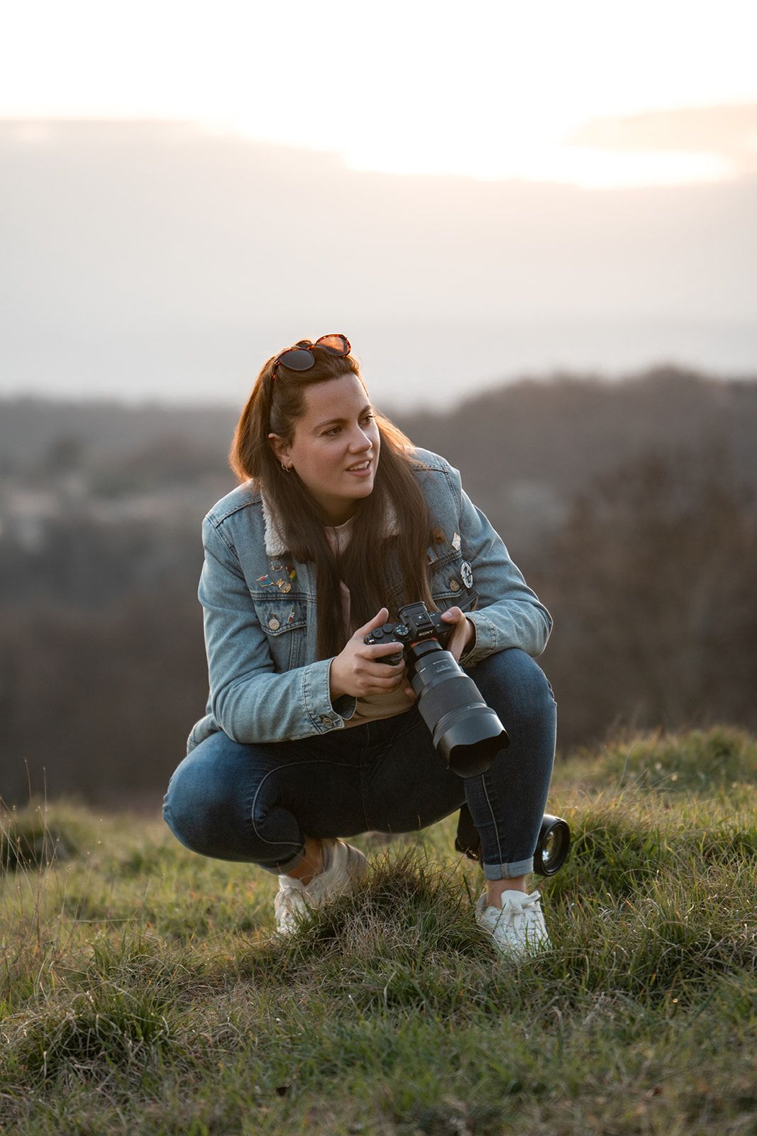 Smiling woman with camera crouching on a grassy hill at golden hour sunset.