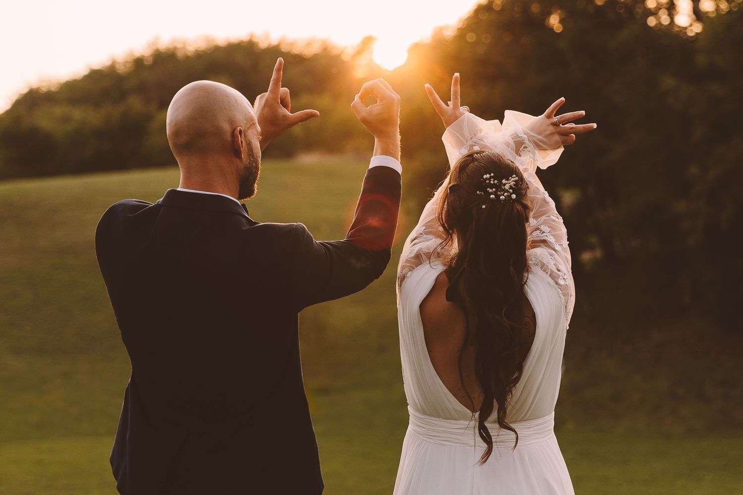 Bride and groom make heart shapes with hands raised against glowing sunset on lush green wedding grounds.