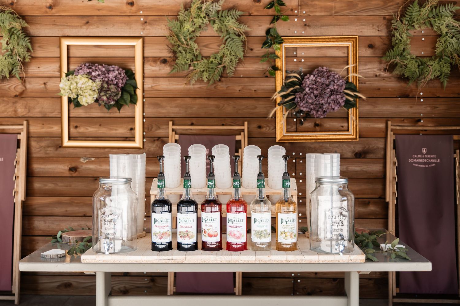 Rustic wooden wall display with gold frames, dried hydrangeas, and syrup bottles arranged on a wedding bar table.