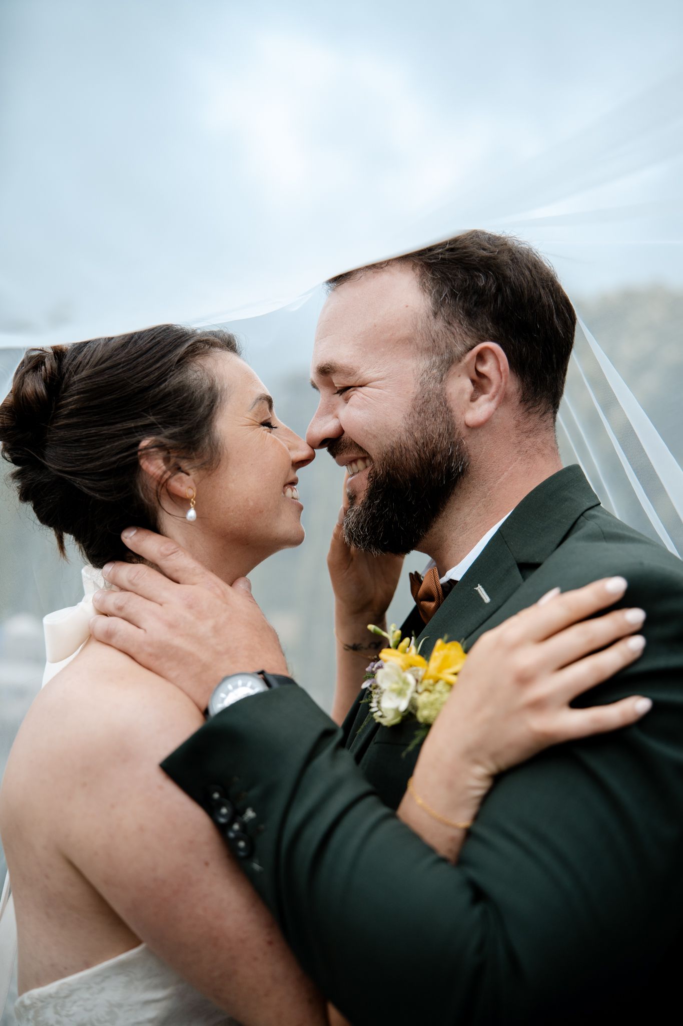 Smiling bride and bearded groom embracing under a veil with yellow floral boutonniere on a cloudy day.
