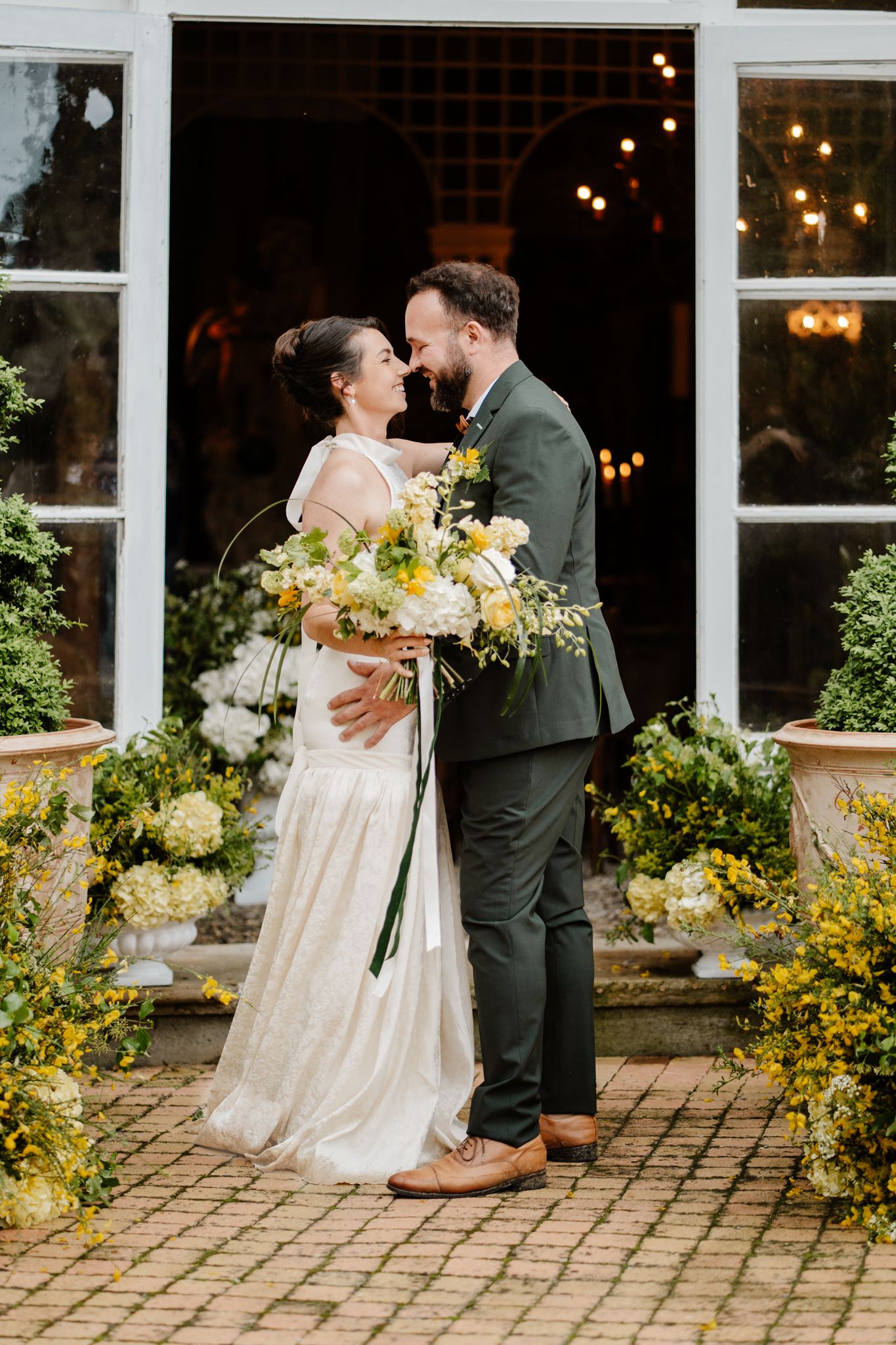 Bride and groom embracing outside venue doors surrounded by white and yellow floral arrangements.