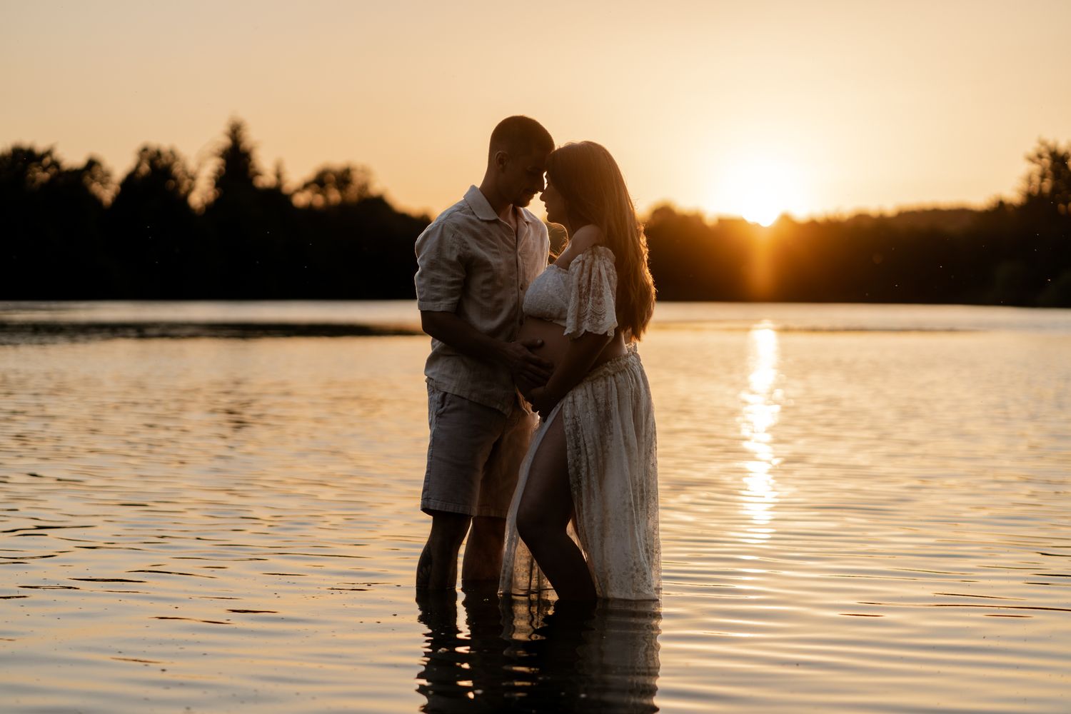 Couple sharing intimate moment standing in lake water during warm golden sunset, woman in white dress.