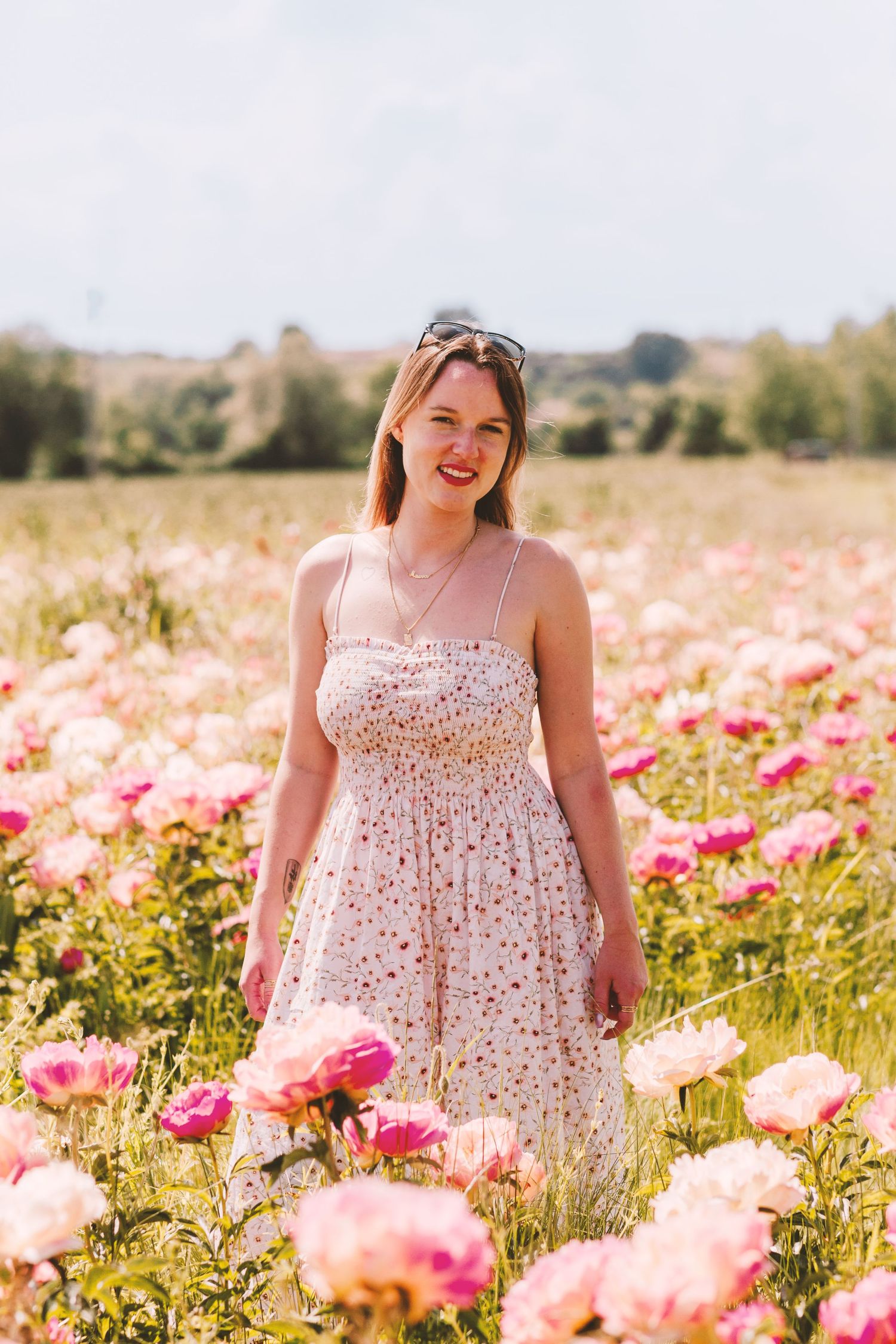 Smiling woman in floral sundress standing in a vibrant pink peony flower field on a sunny day.