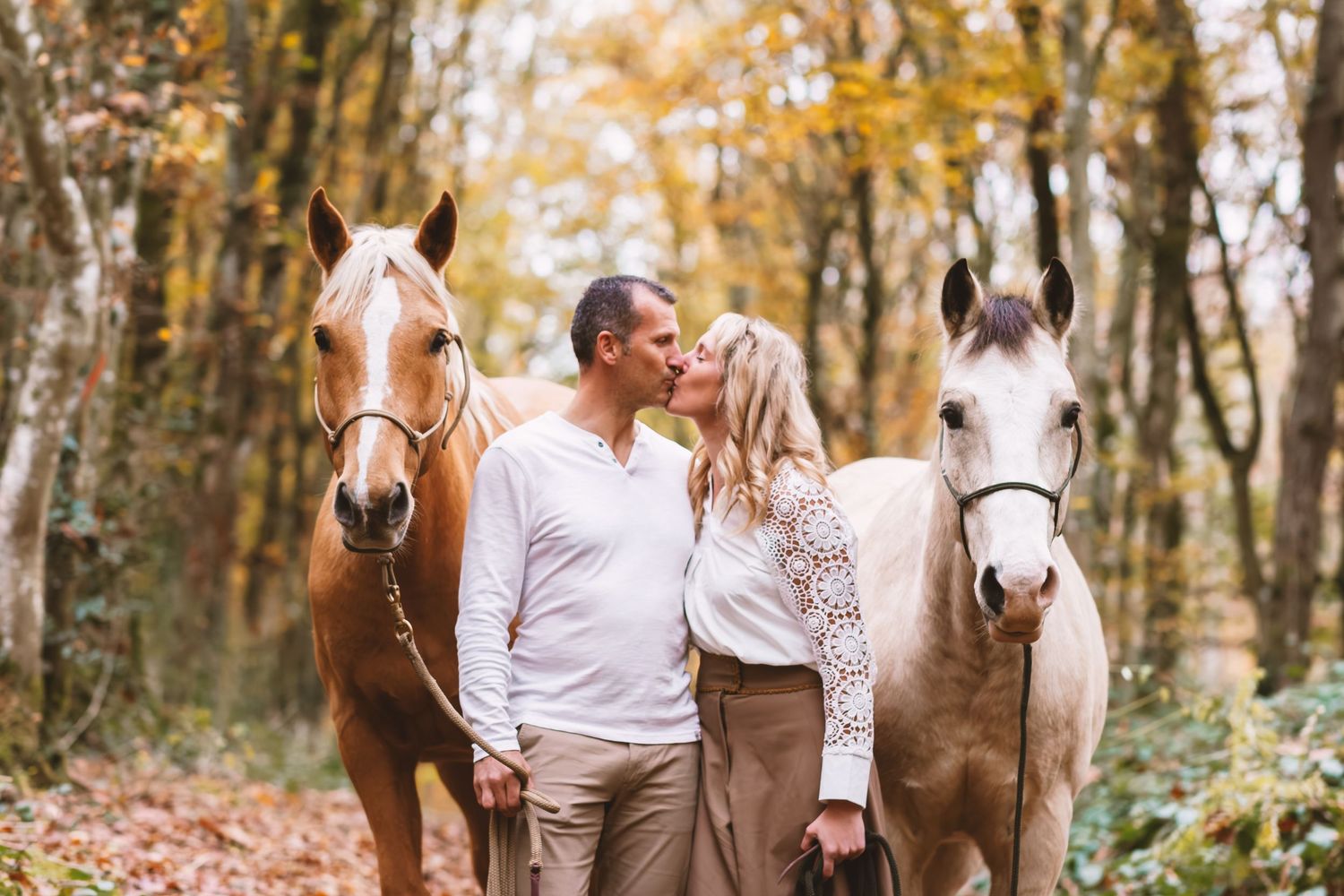 Couple kissing between two horses on an autumn forest path with golden foliage.